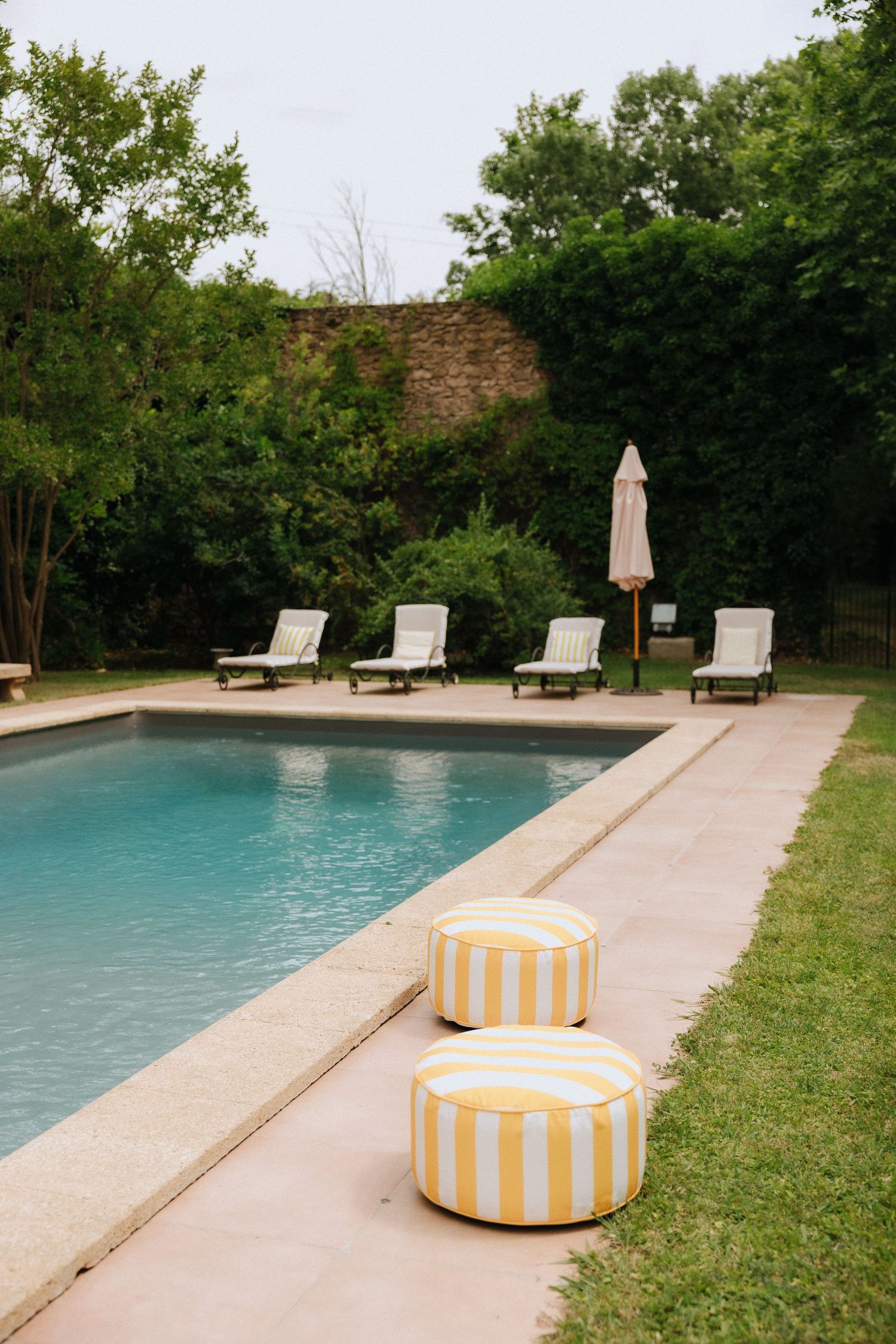 Poolside lounge area with striped yellow ottomans and chaise lounges against stone wall.