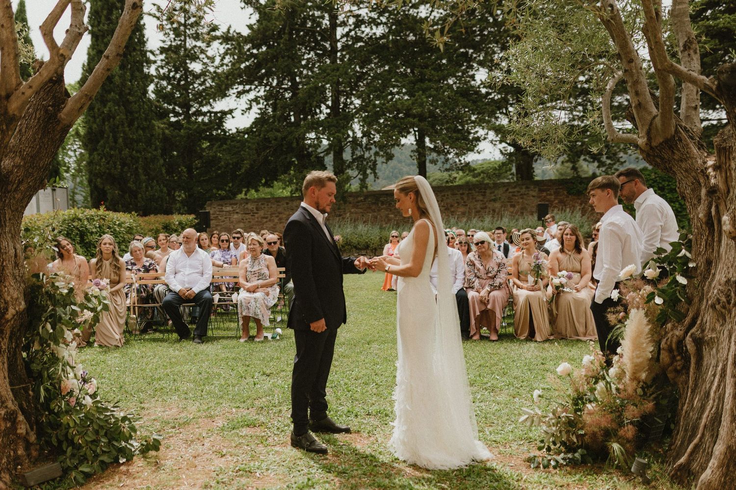 Outdoor wedding ceremony taking place between trees with guests seated on wooden chairs in a garden setting.