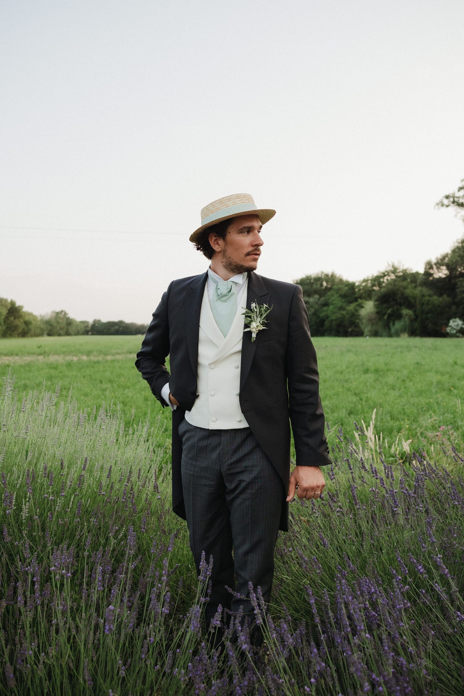 Figure in a dark suit and wide-brimmed hat walking through a lavender field on an overcast day.