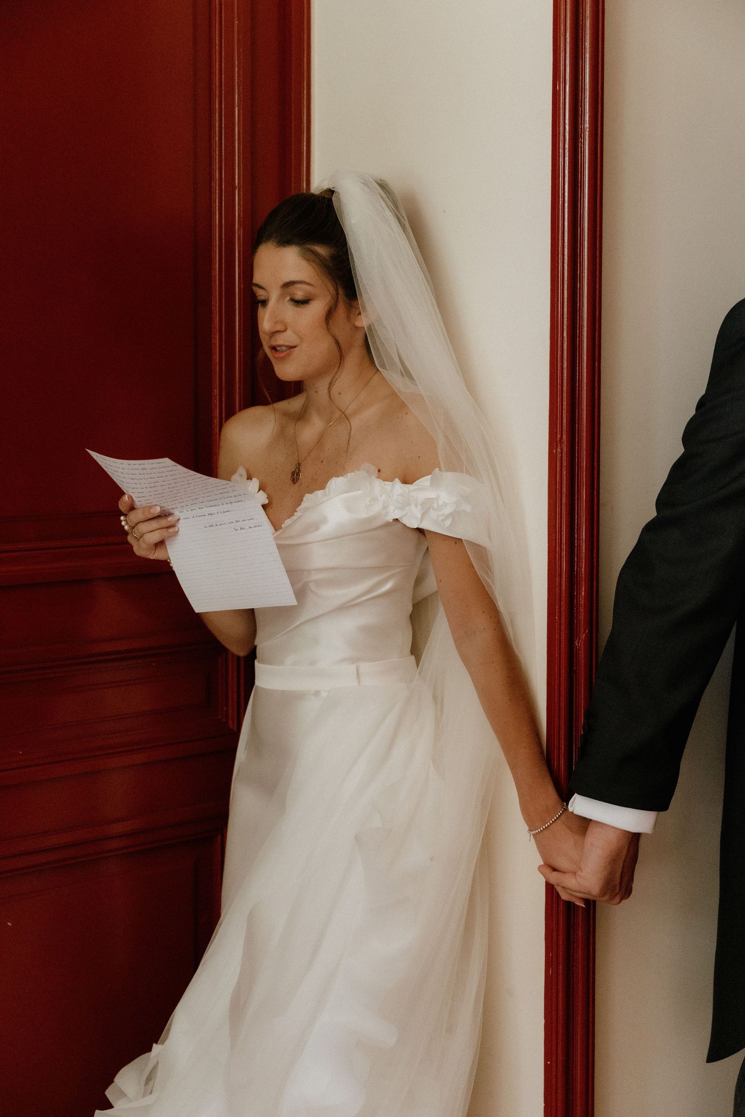 A bride in white dress reads a letter while standing against a red wall.