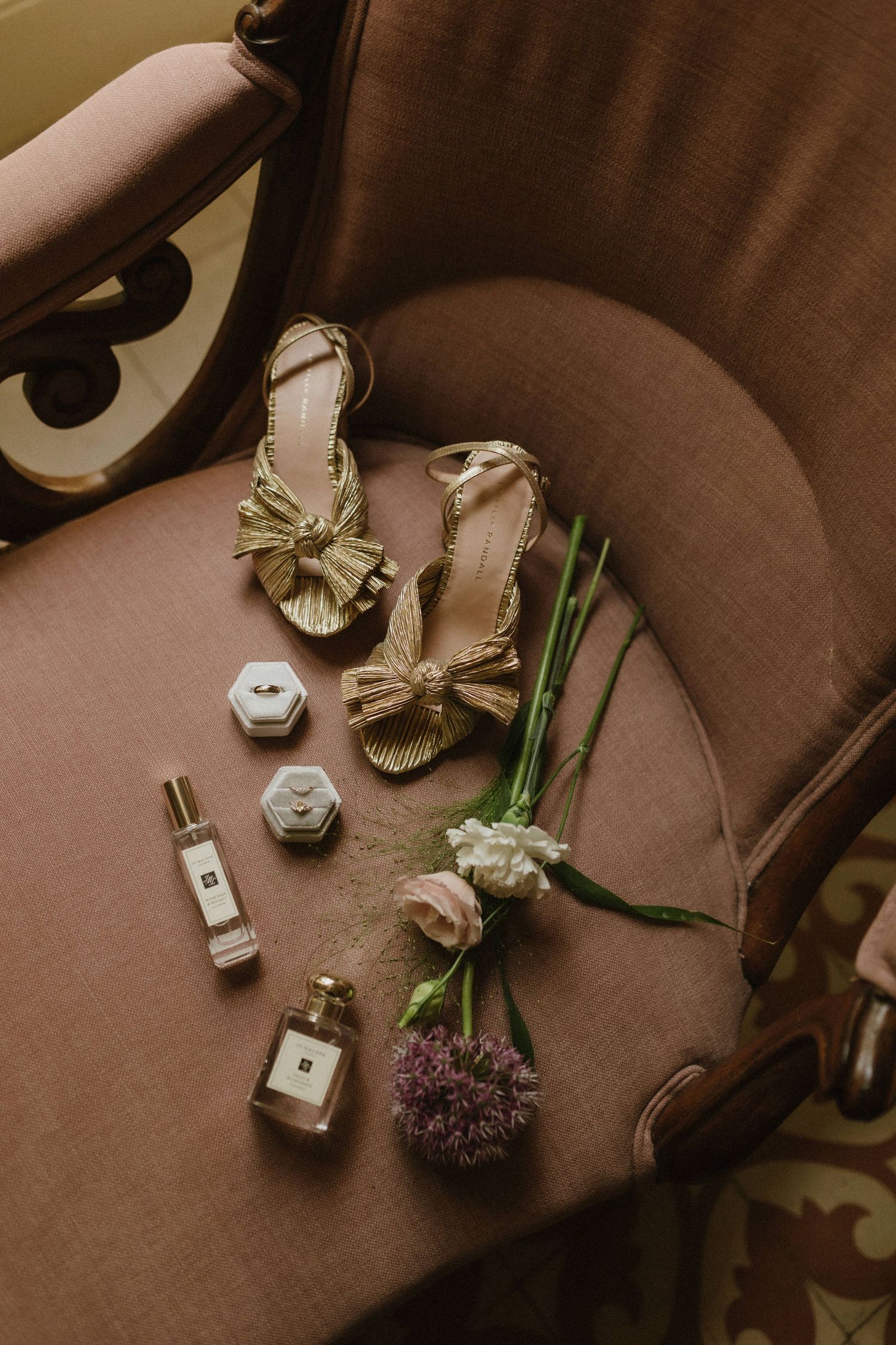 Elegant gold bow sandals arranged with perfume bottles and fresh flowers on a brown velvet chair.