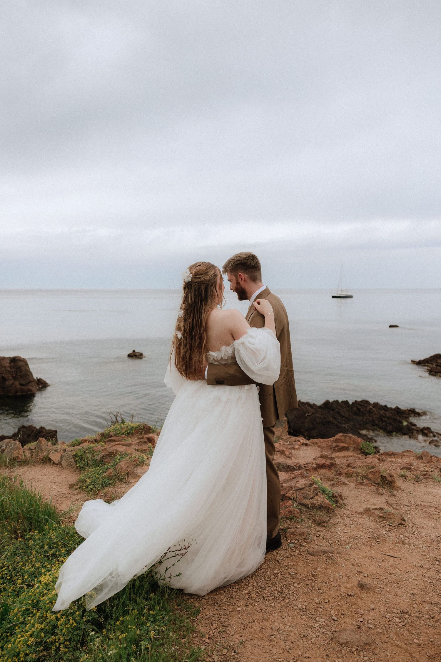 A romantic couple in wedding attire embrace on a coastal cliff overlooking a misty ocean on their special day.