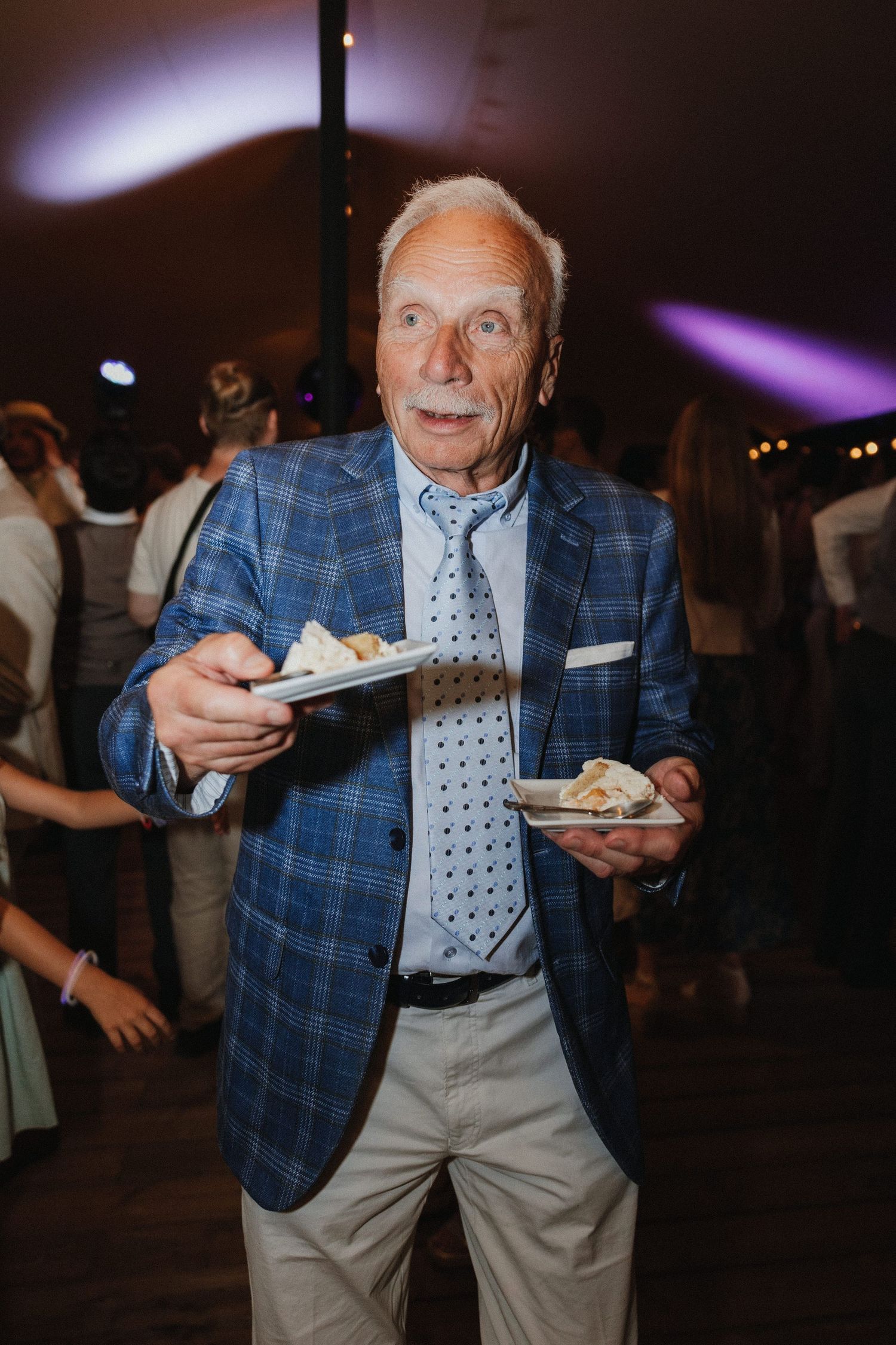 A guest in a blue plaid blazer and tie enjoys dessert plates at an evening celebration event.