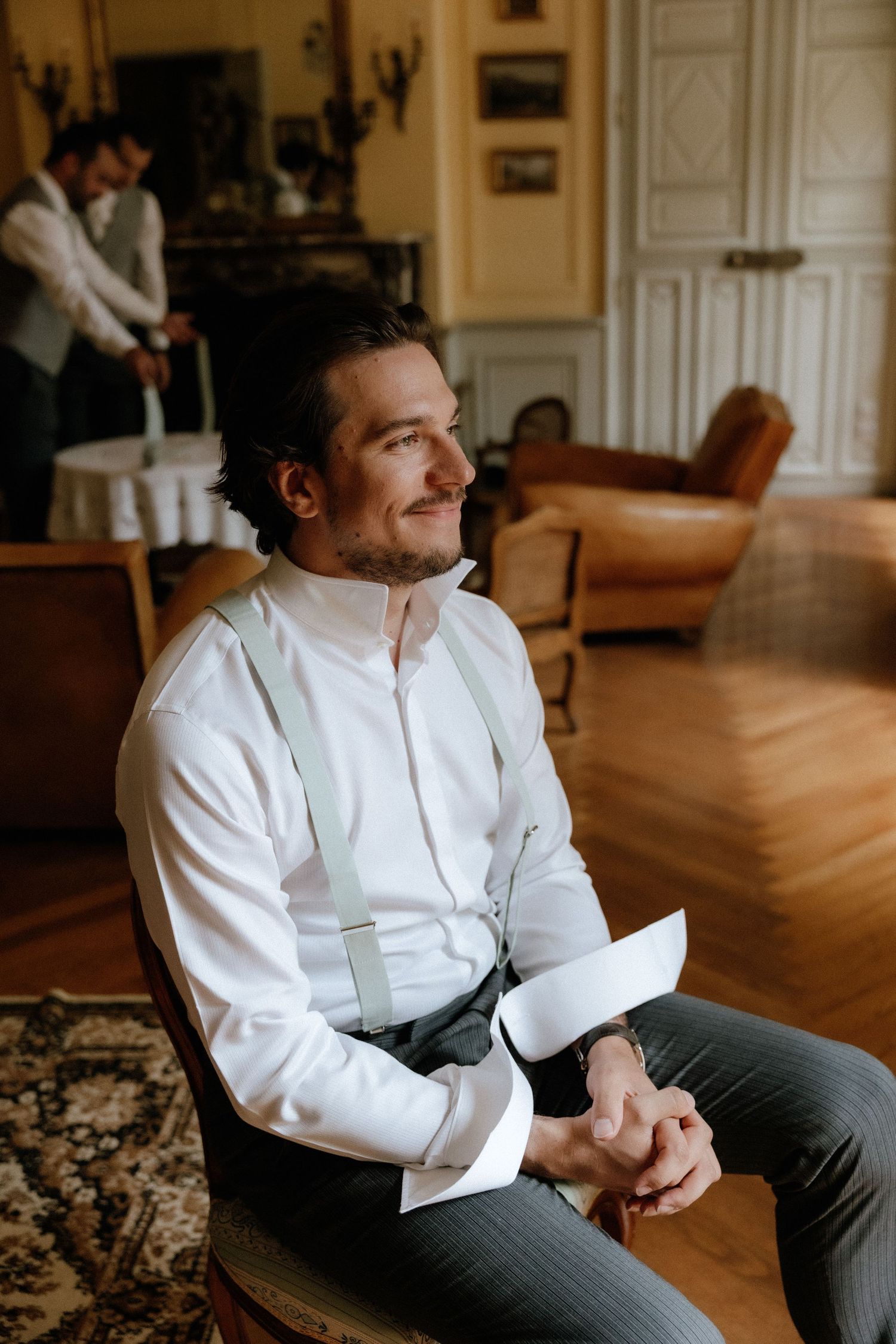 Person in white dress shirt and gray pants sits thoughtfully in an elegant room with wooden floors.