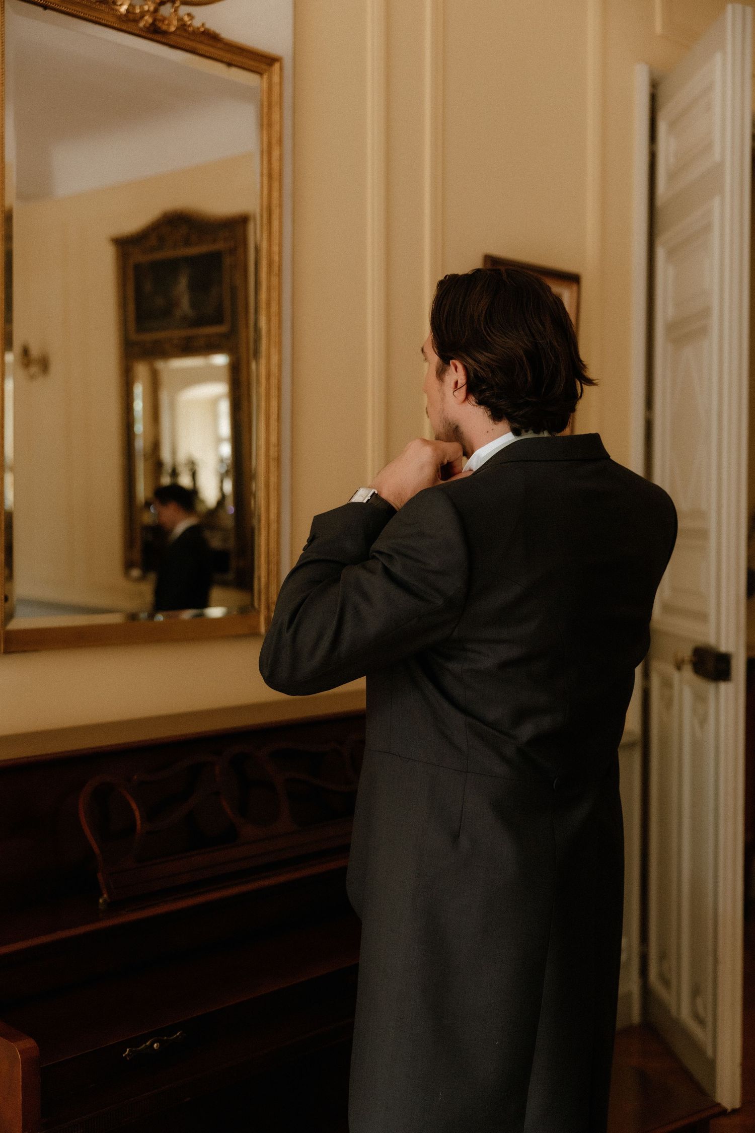 Person in formal attire adjusting collar while looking in a gold-framed mirror.