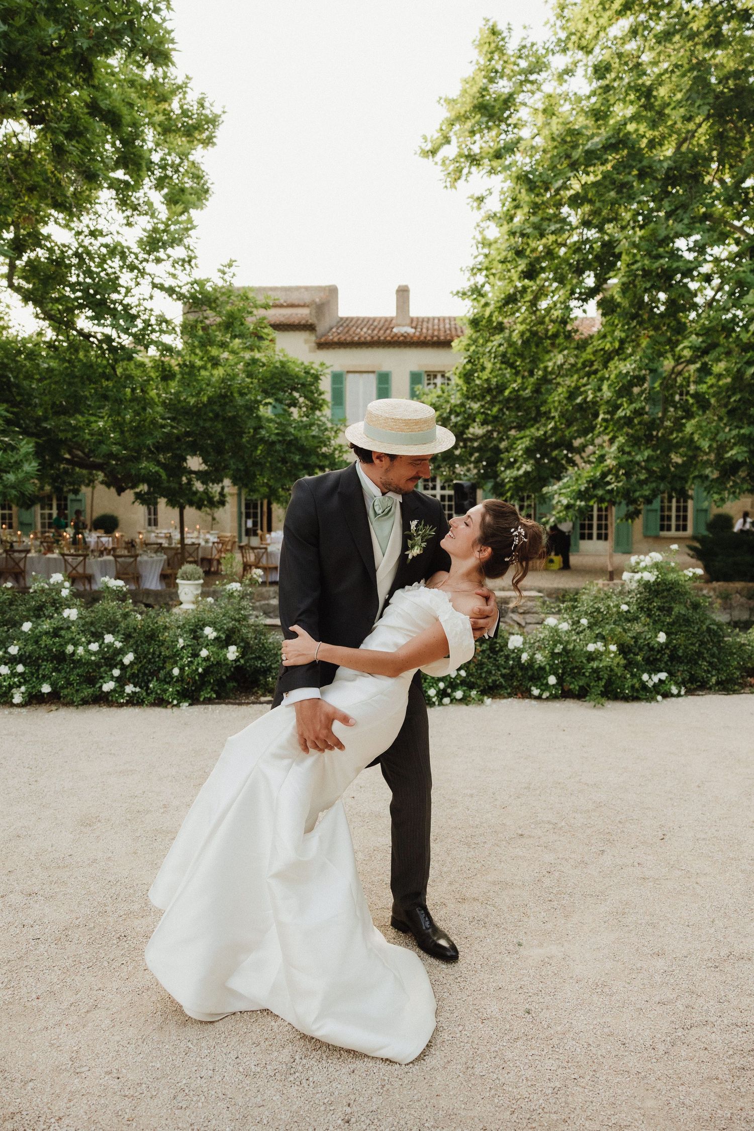 A couple in formal wedding attire performs a romantic dip pose on a sandy outdoor venue surrounded by trees.