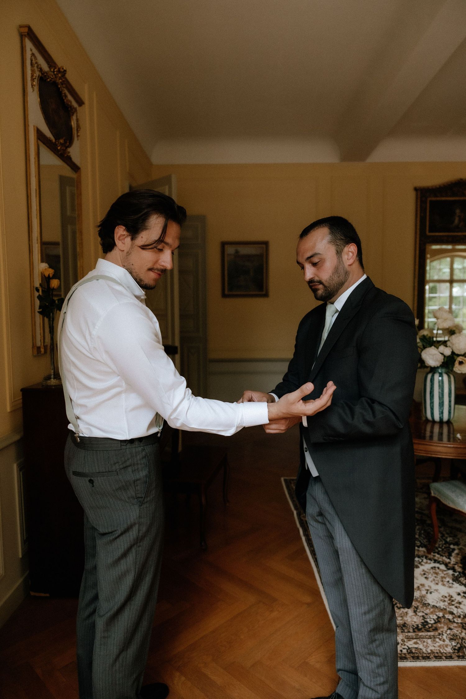 Two men in formal attire examining something in an elegant room with hardwood floors.