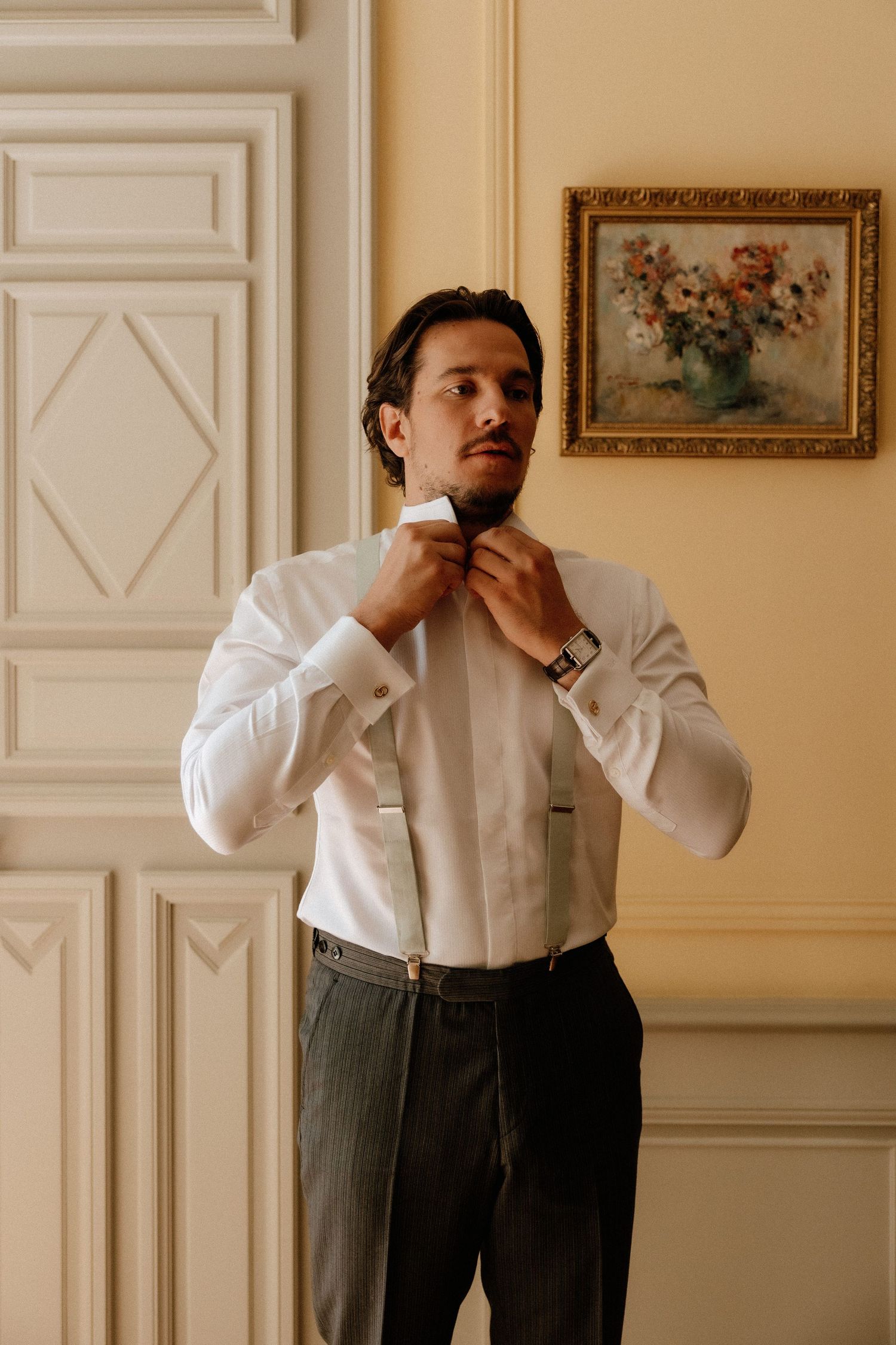 Groom adjusting his tie while getting ready in an elegant room with ornate wall decor.
