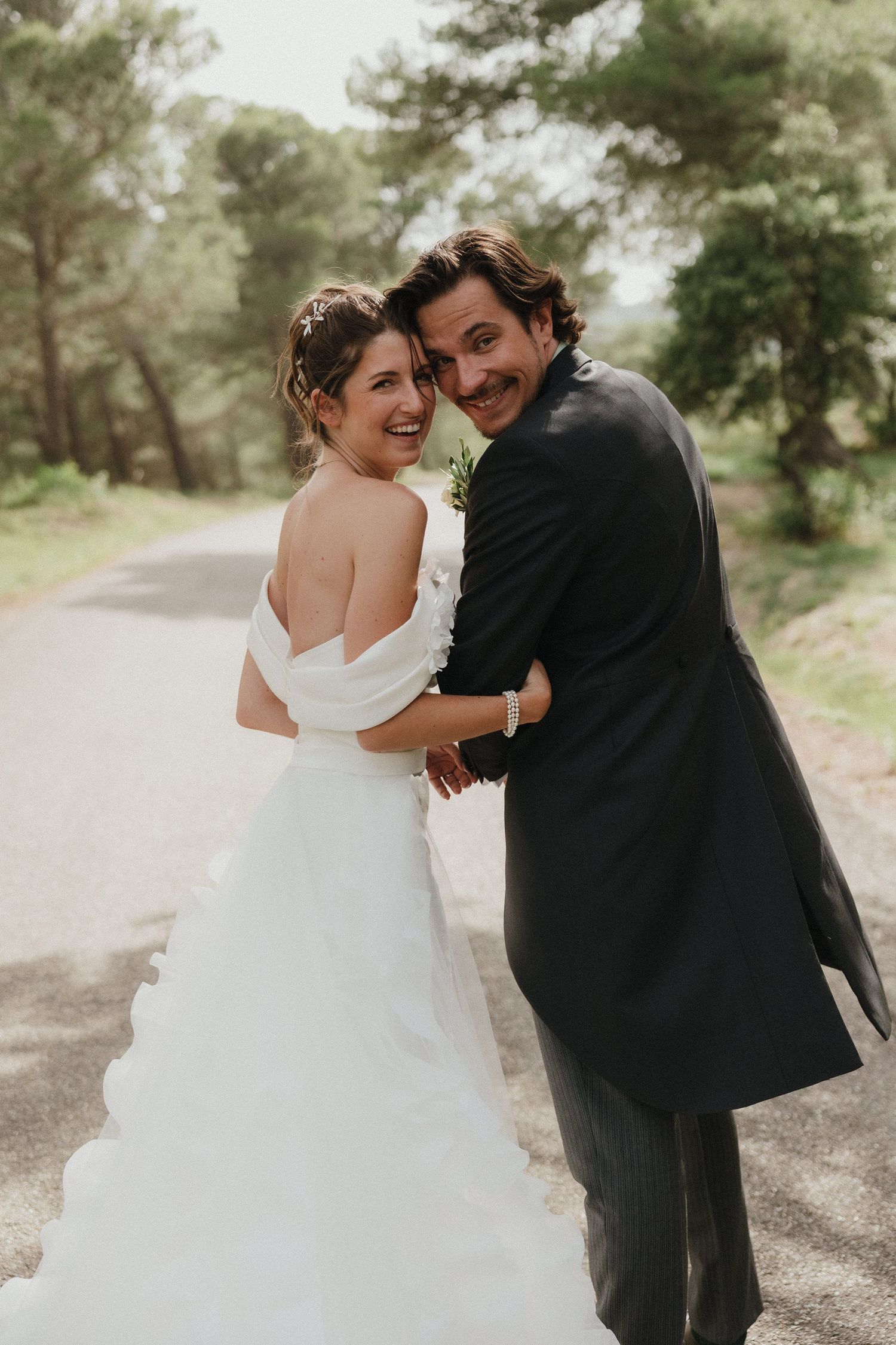 Wedding couple embraces and laughs together on a tree-lined path.