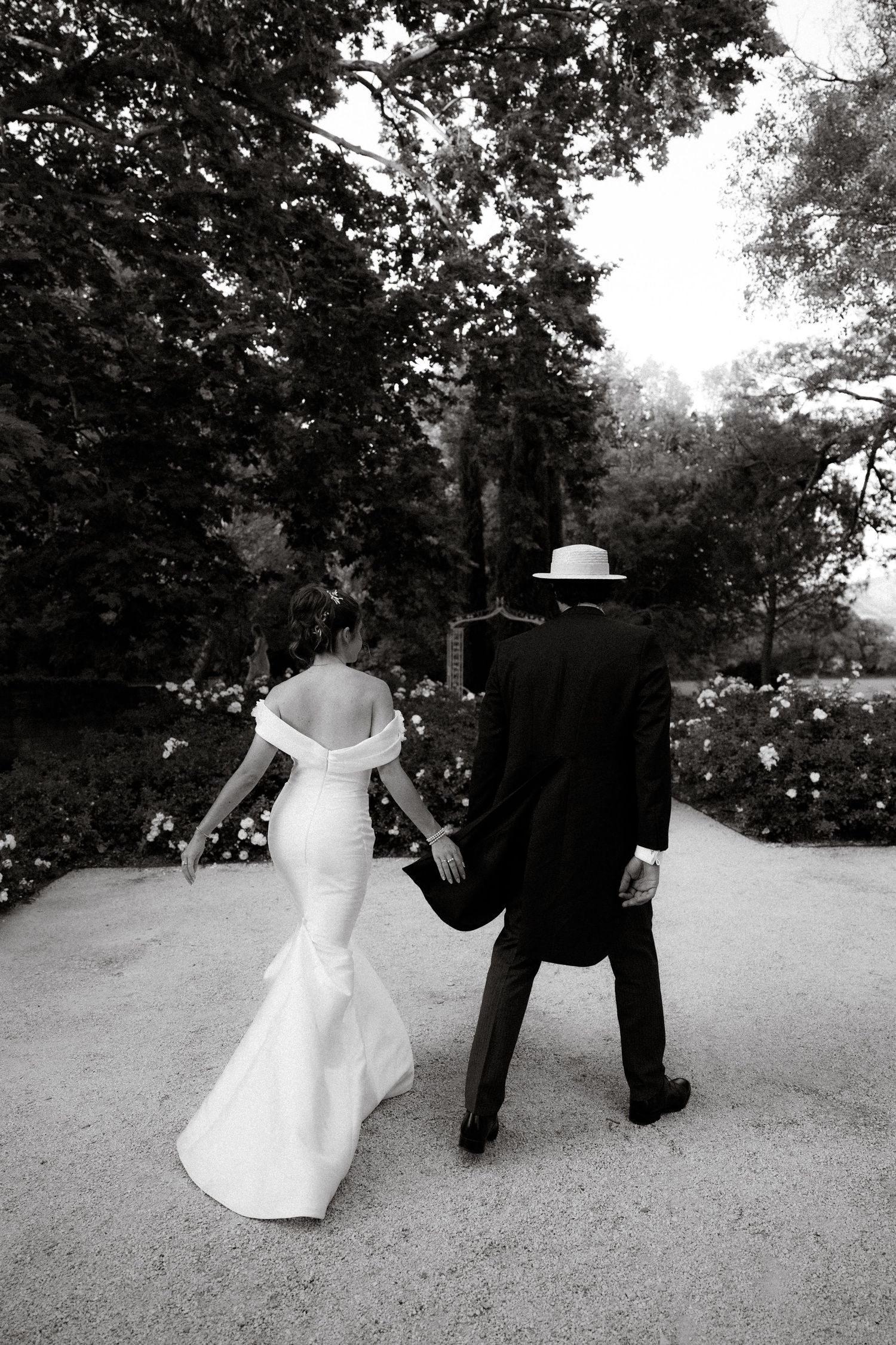A bride and groom walk hand in hand down a garden path in black and white.
