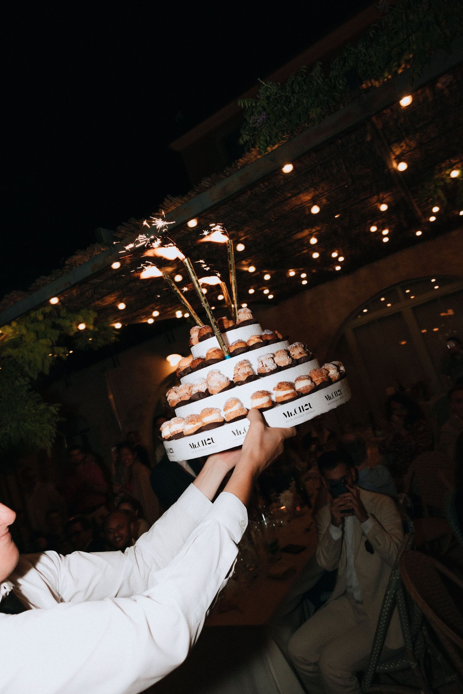 A tiered wedding cake being held up with sparklers and twinkling lights in the background at an evening reception.