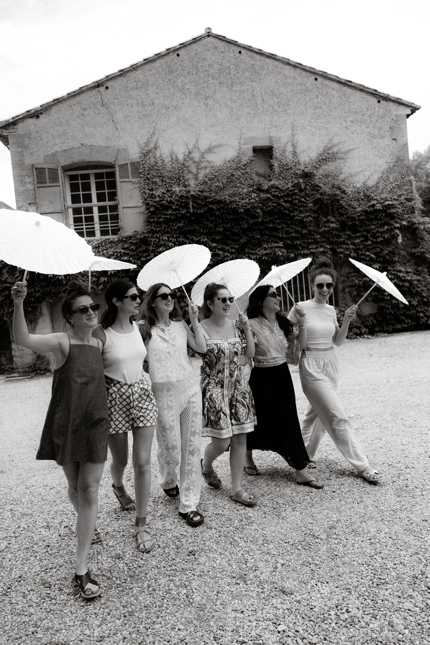 A group of friends pose with white parasols in front of a rustic building during a sunny outdoor gathering.