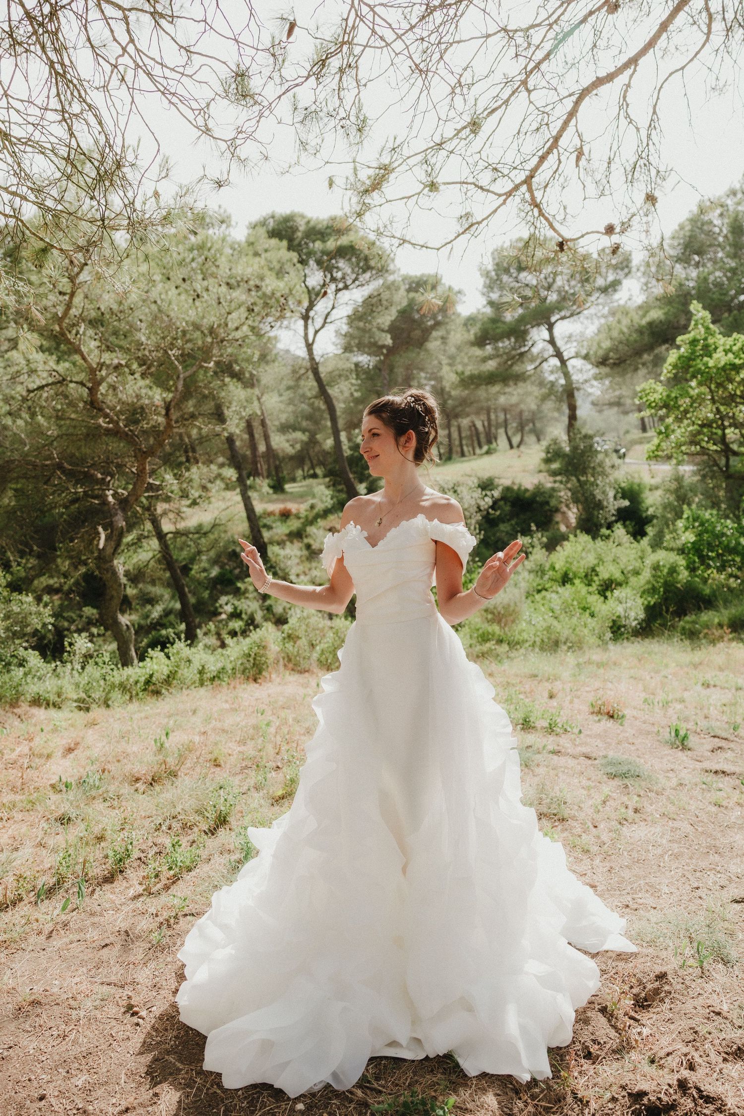 Bride in flowing white dress twirling outdoors in natural sunlight among trees.