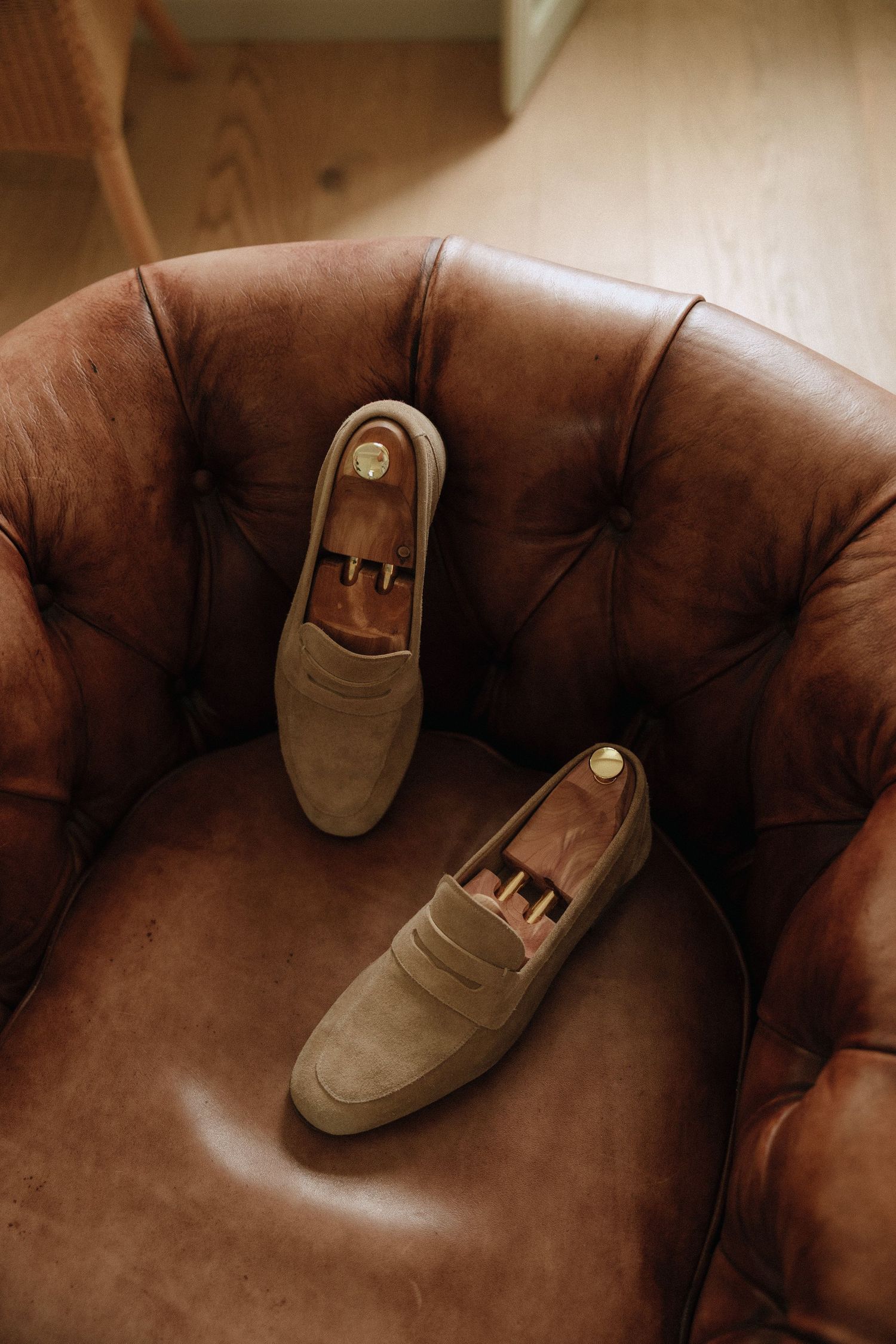 A pair of tan suede loafers resting on a brown leather tufted chair captured in warm lighting.