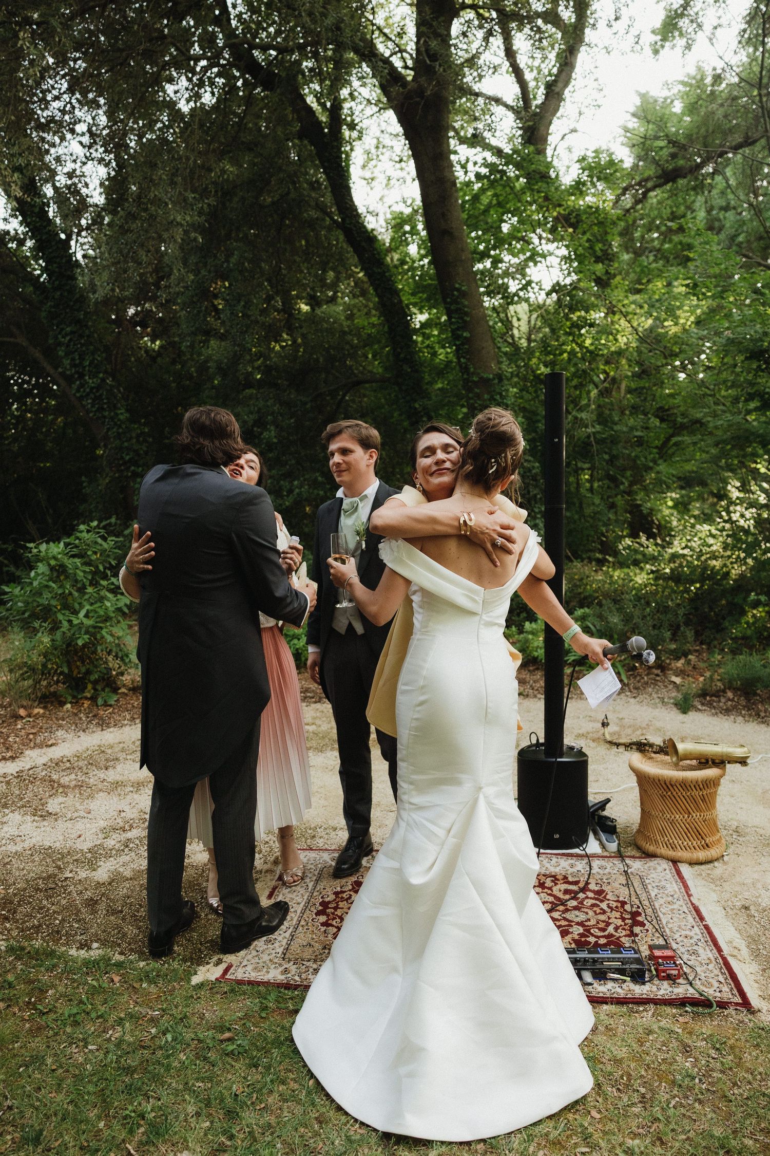 Wedding celebration in a garden setting with guests gathering on outdoor rugs under trees.