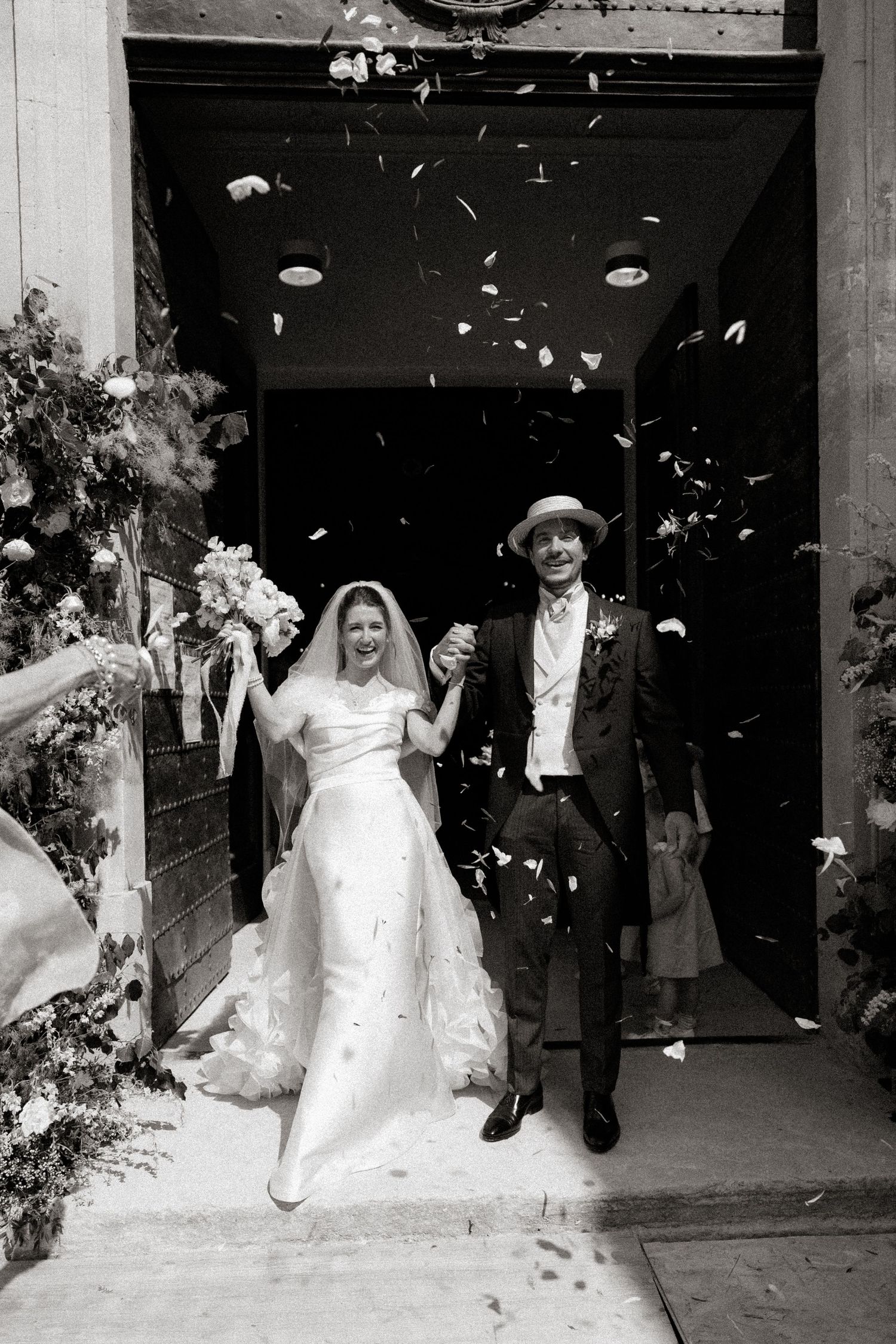 A black and white photo of a newlywed couple exiting a church while flower petals fall around them.
