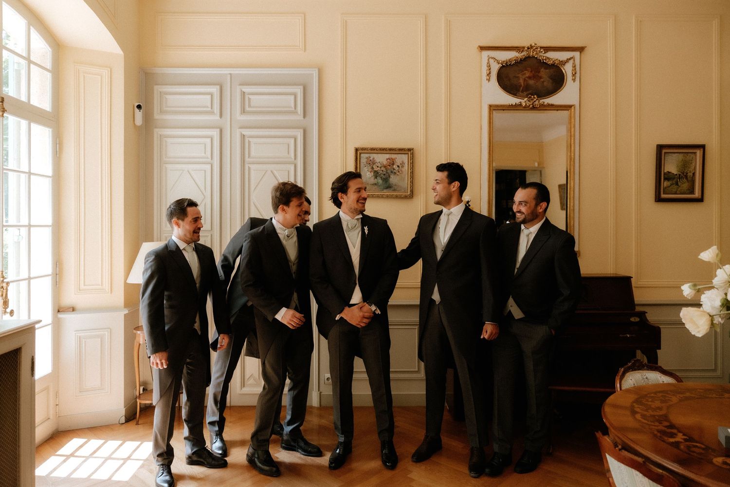 A group of men in dark suits stand in an elegant white-paneled room during a formal gathering.