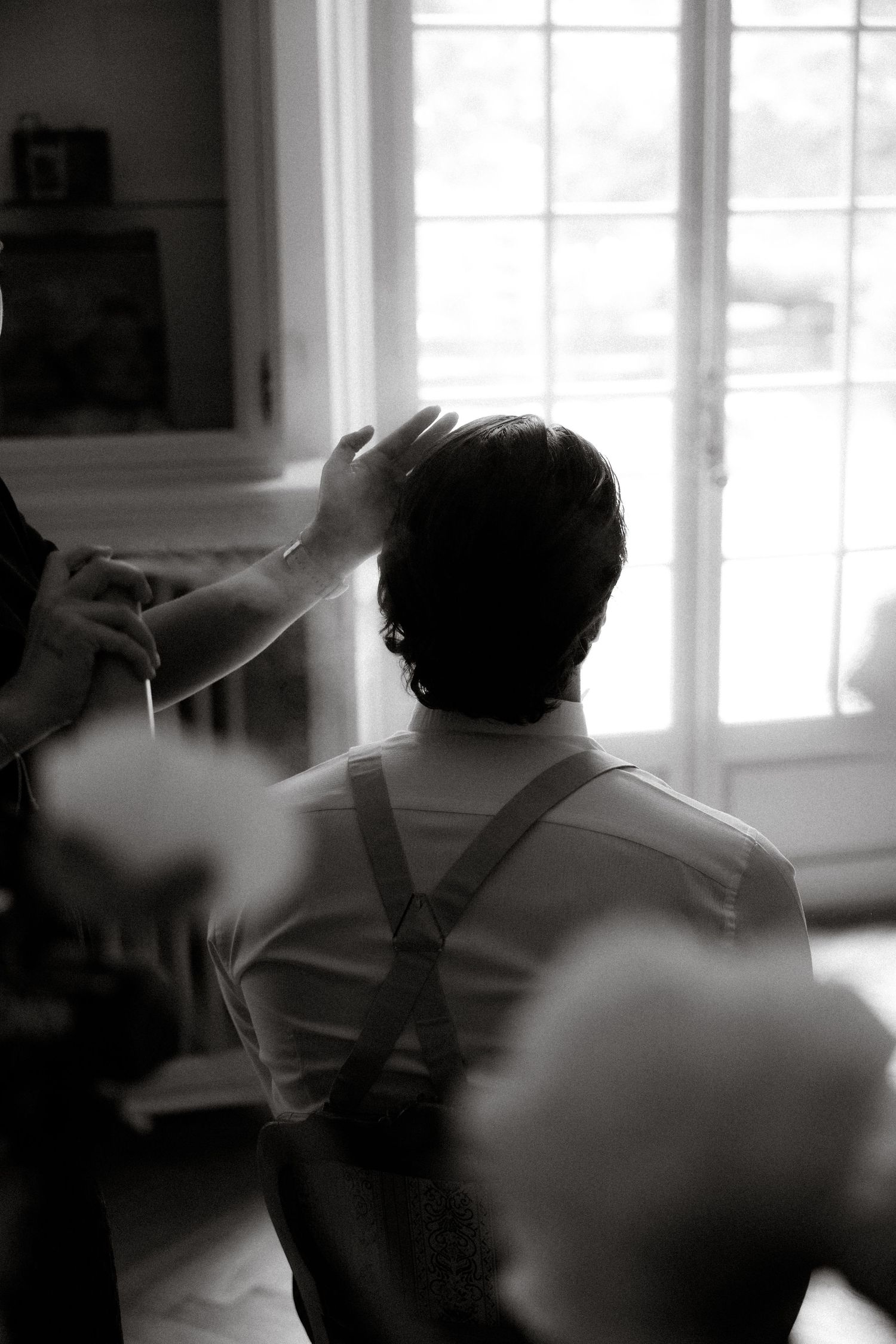 A black and white photo showing someone getting their hair styled near a bright window.