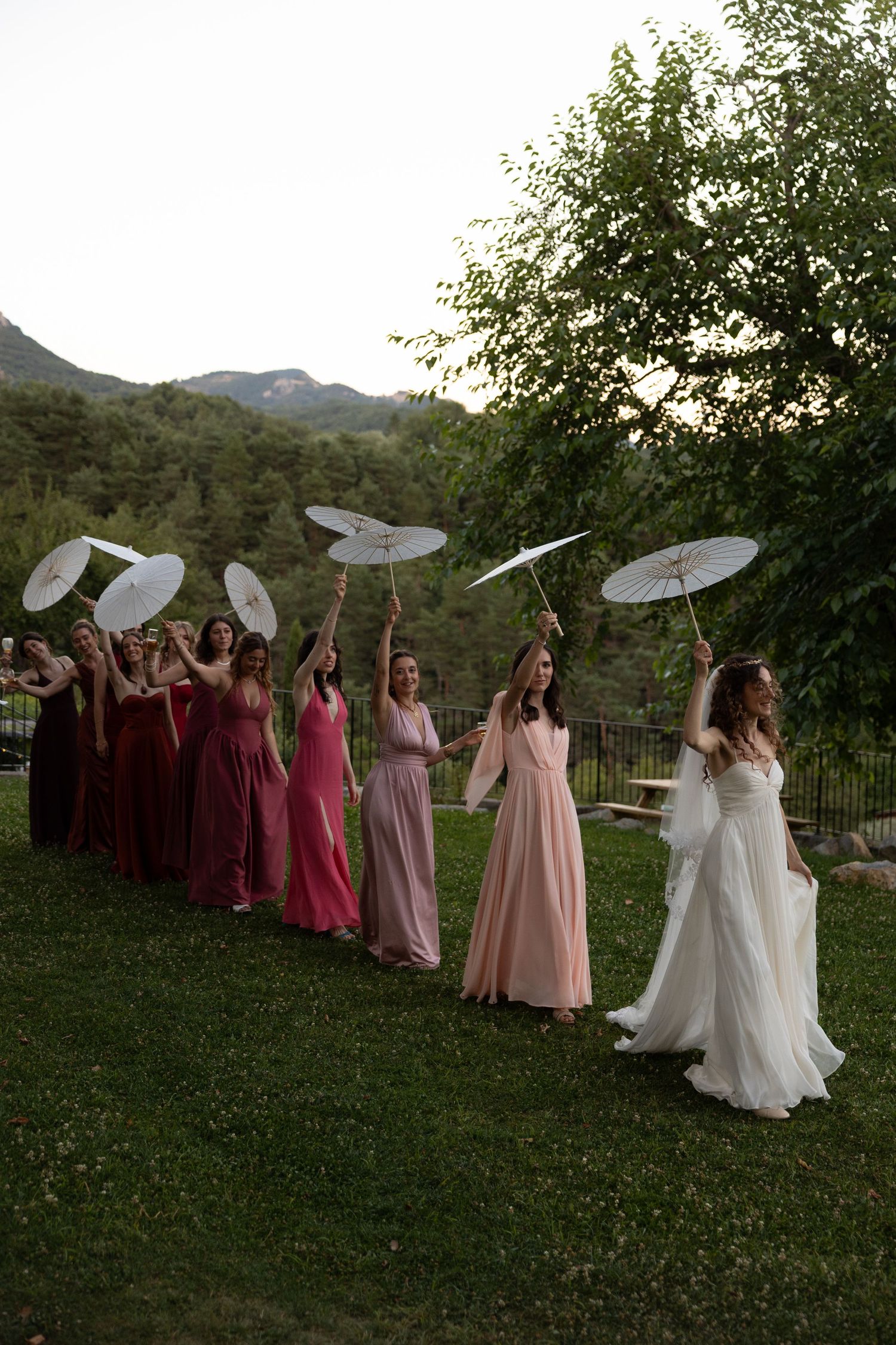 Bridal party holding white parasols stands in line on grassy lawn with mountain backdrop at sunset.