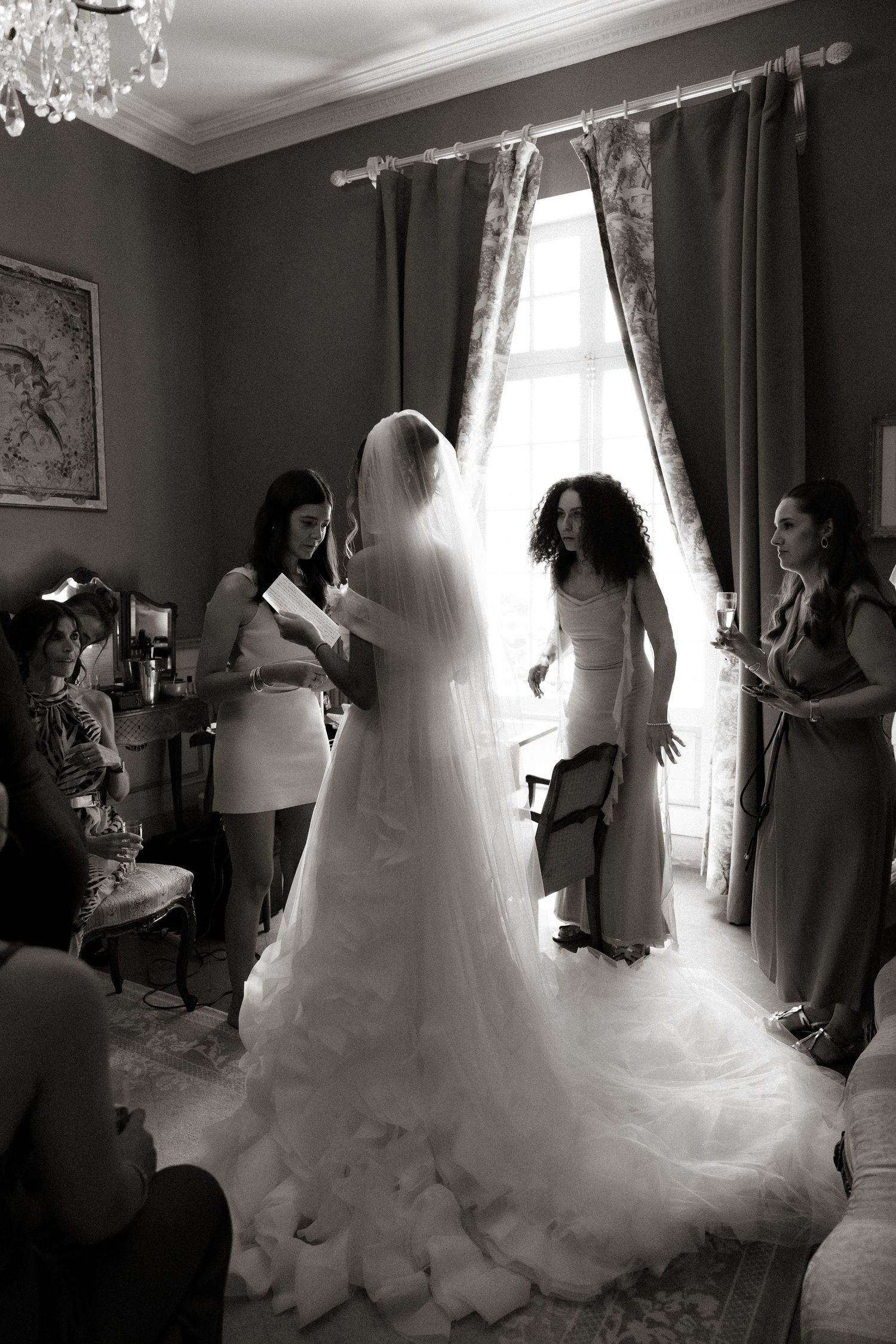 A wedding dress preparation scene showing a flowing white gown being adjusted in an elegant room with curtains.