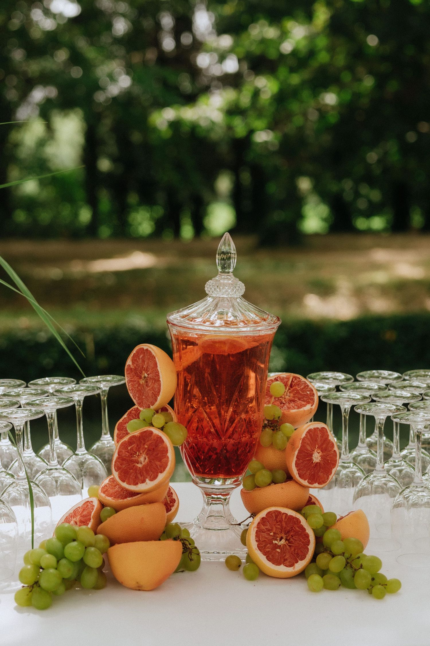 A crystal punch bowl with red grapefruit punch surrounded by fresh citrus fruits and green grapes on a white table.
