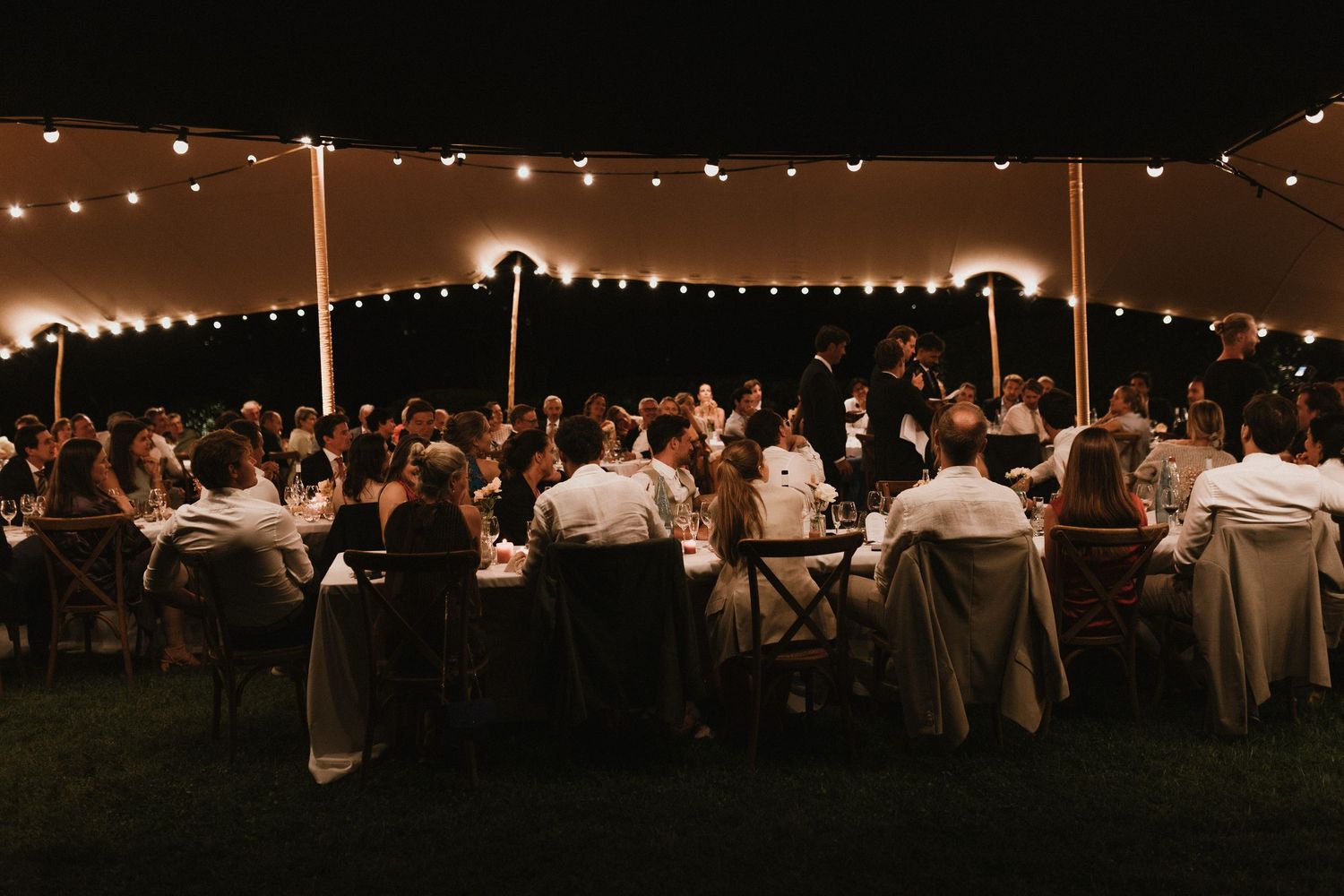 Wedding reception dinner under soft string lights and tent at dusk with guests seated at long tables.