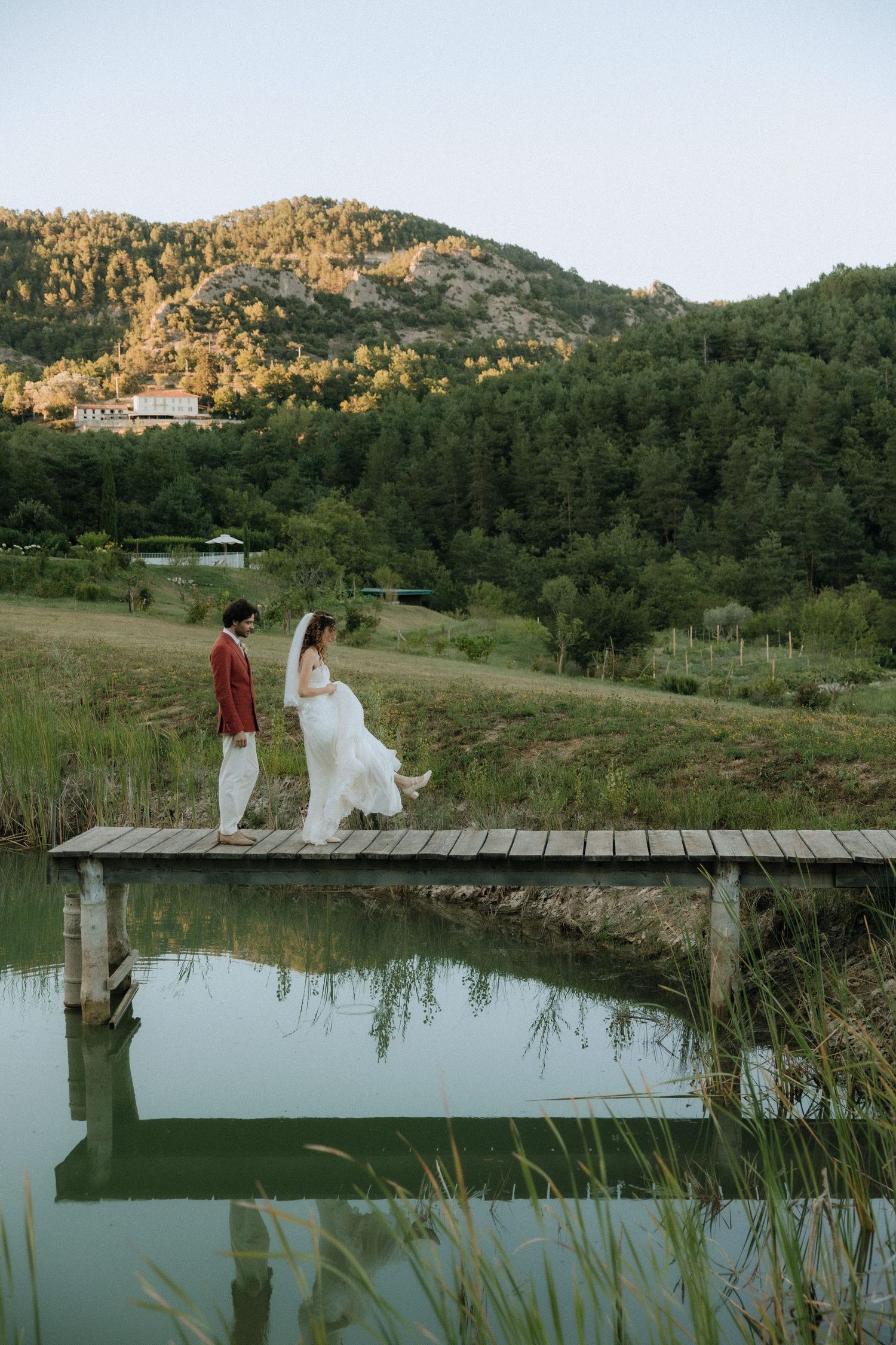 A couple walks along a wooden dock over a serene lake surrounded by mountain scenery at sunset.