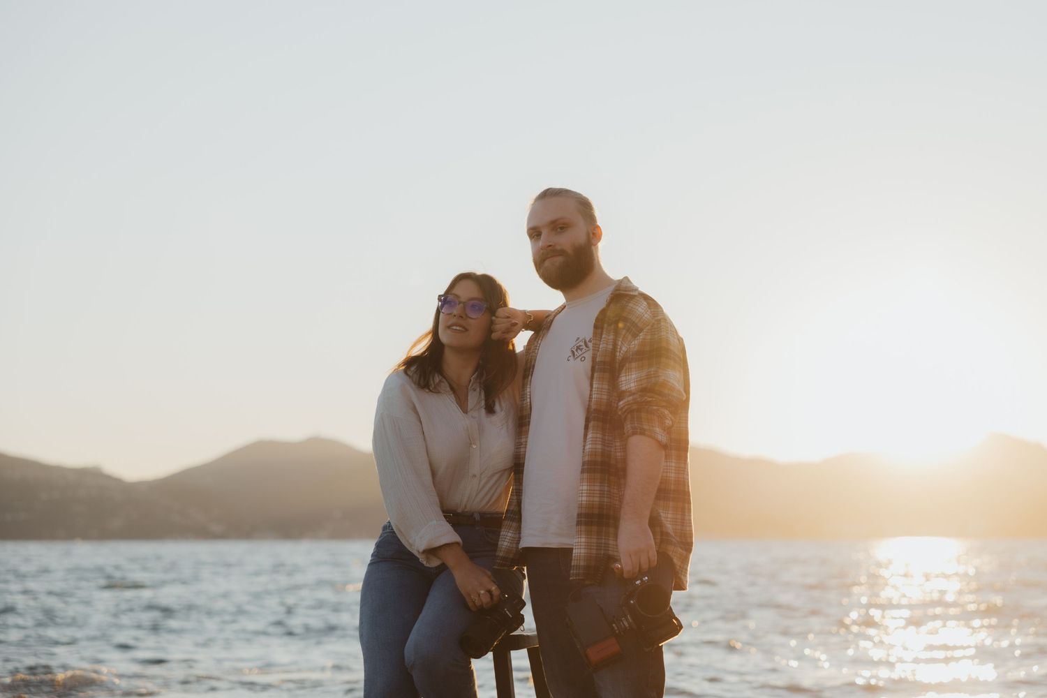 A couple embraces on a rocky beach at sunset with mountains and water in the background.