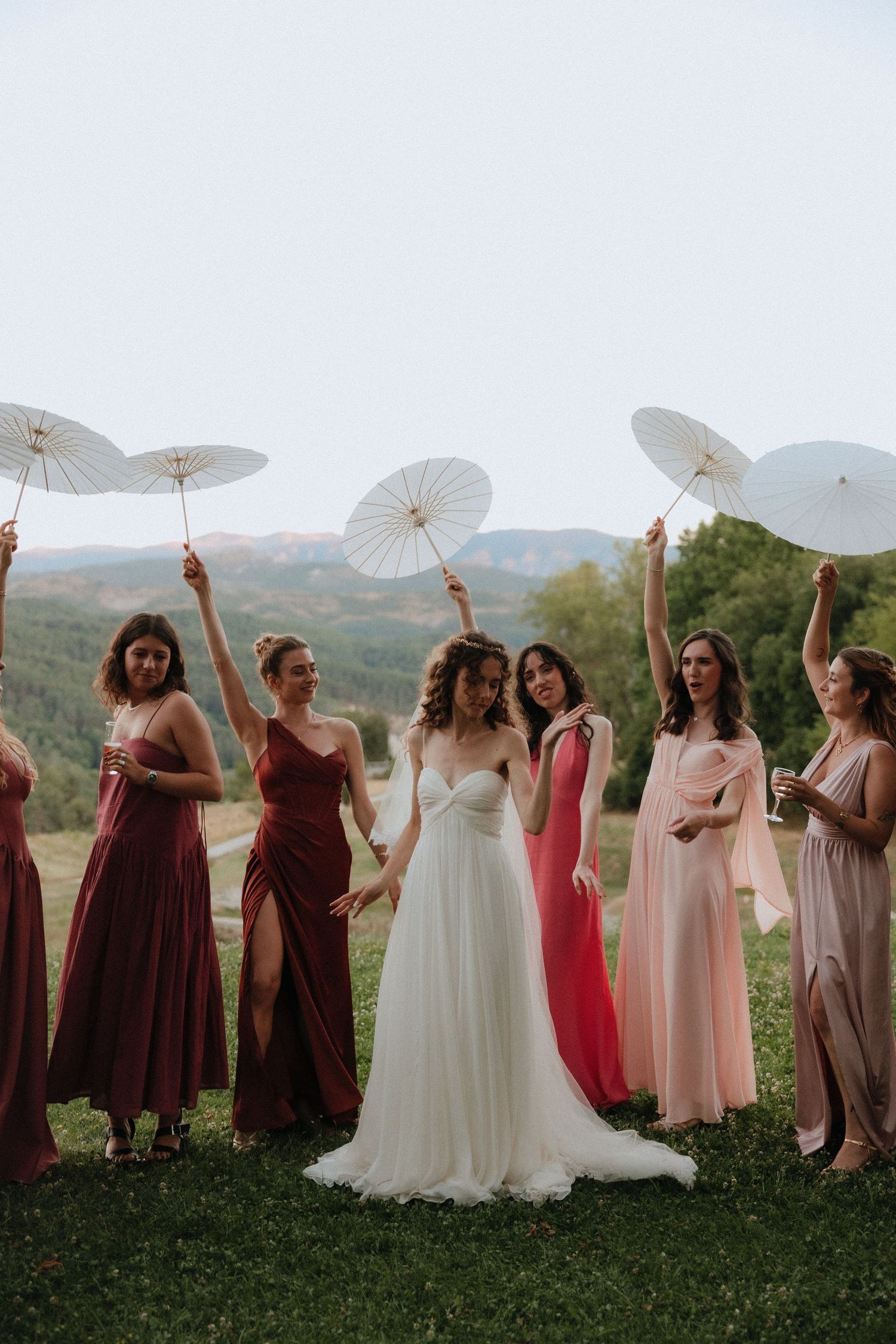 Wedding party poses with white parasols on a grassy hilltop with mountains in the distance.