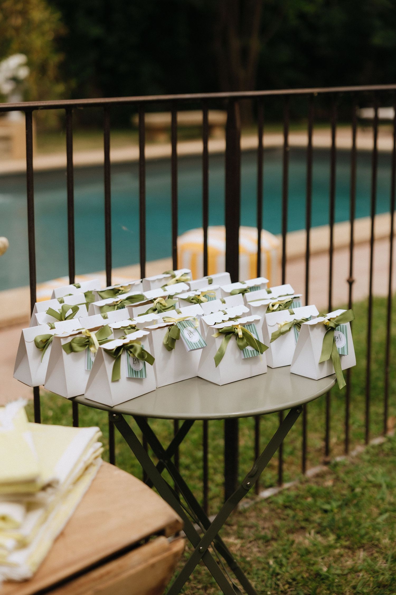 White wedding favor boxes with green ribbons displayed on folding tables near a pool.