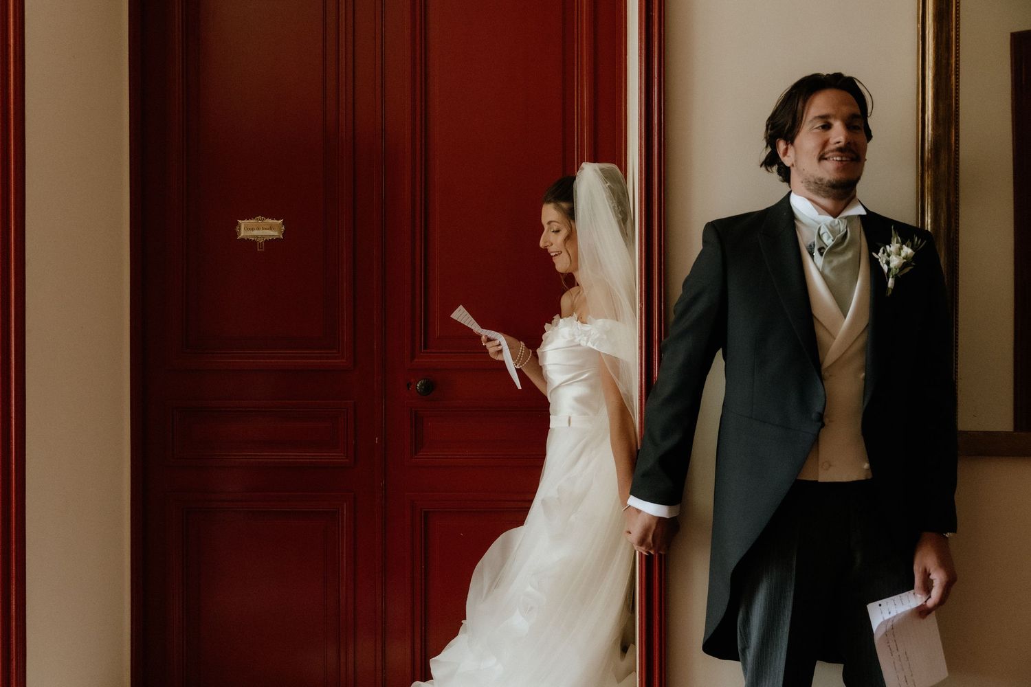 A couple in formal wedding attire stands in an elegant hallway with dark wood doors and red walls.