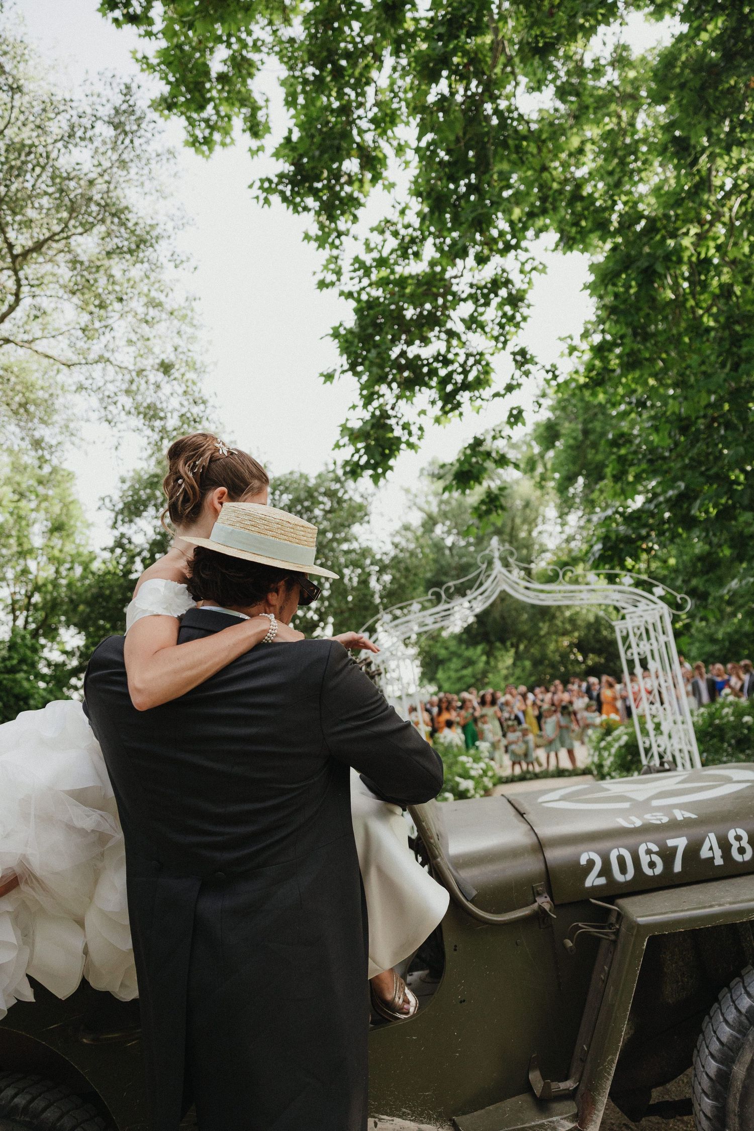 Couple poses with vintage military vehicle during outdoor wedding photoshoot under green trees.