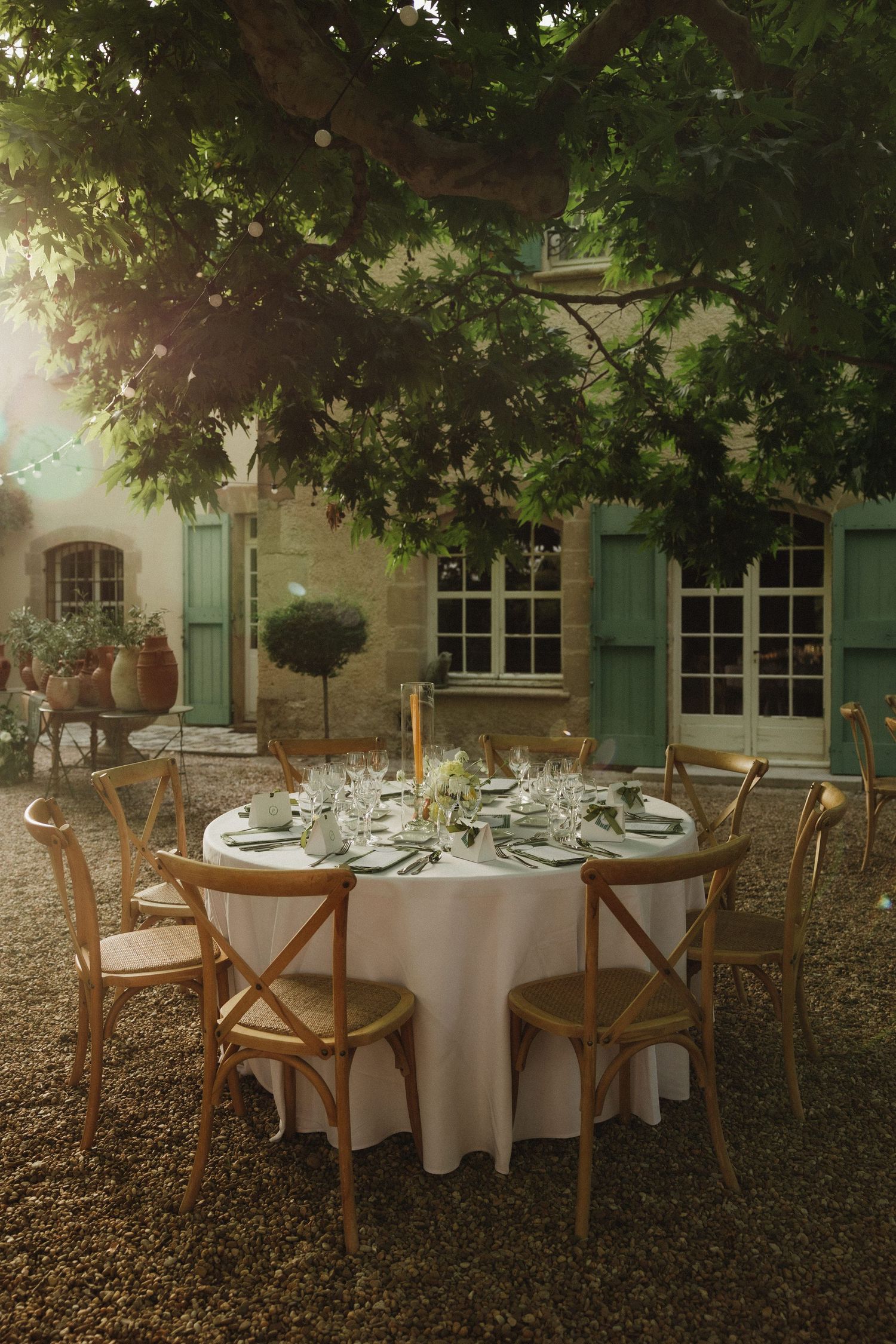 Elegant outdoor wedding reception setup with white tablecloths and wooden chairs under trees.