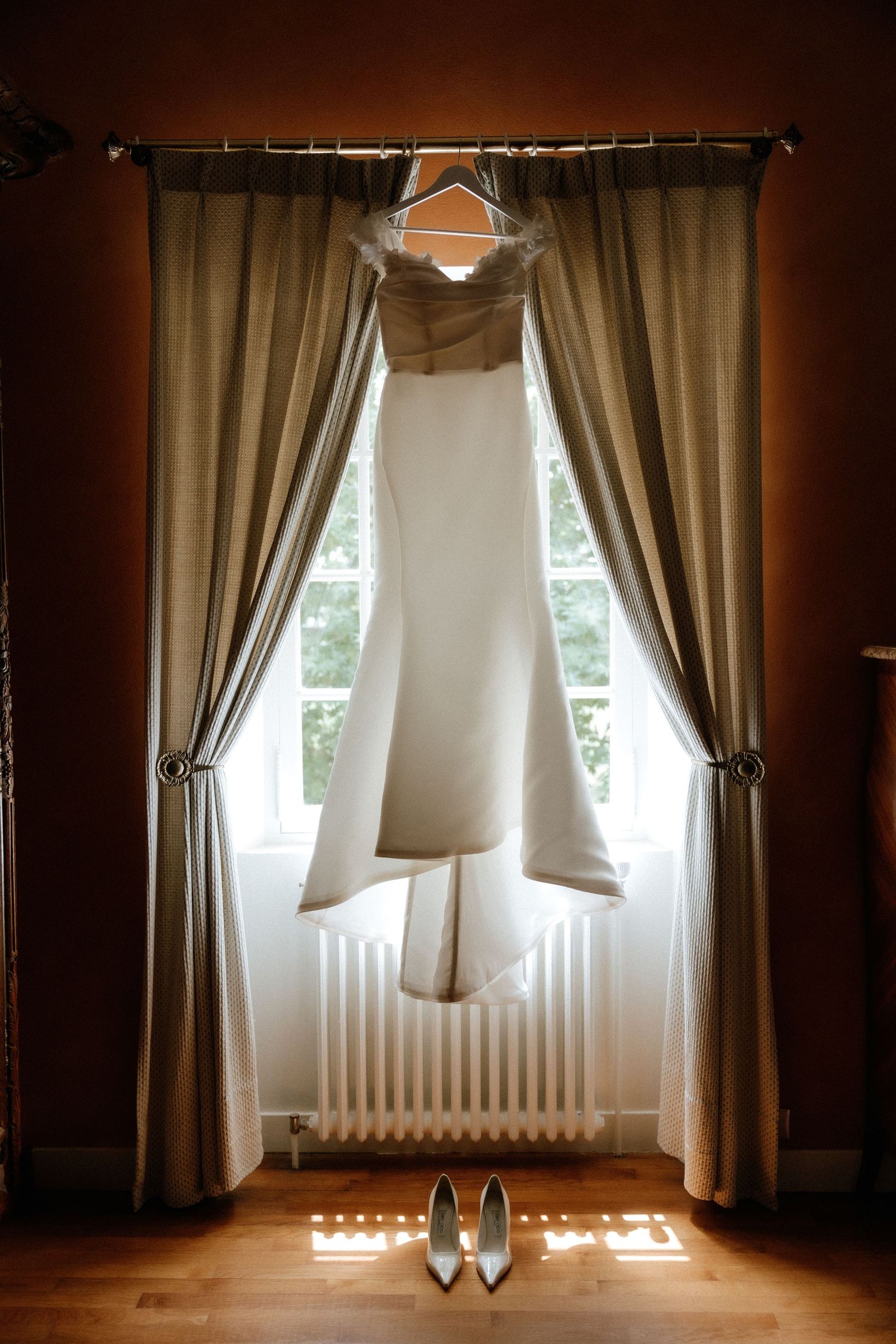 Wedding dress hanging between tan curtains with white heels placed beneath on hardwood floor.