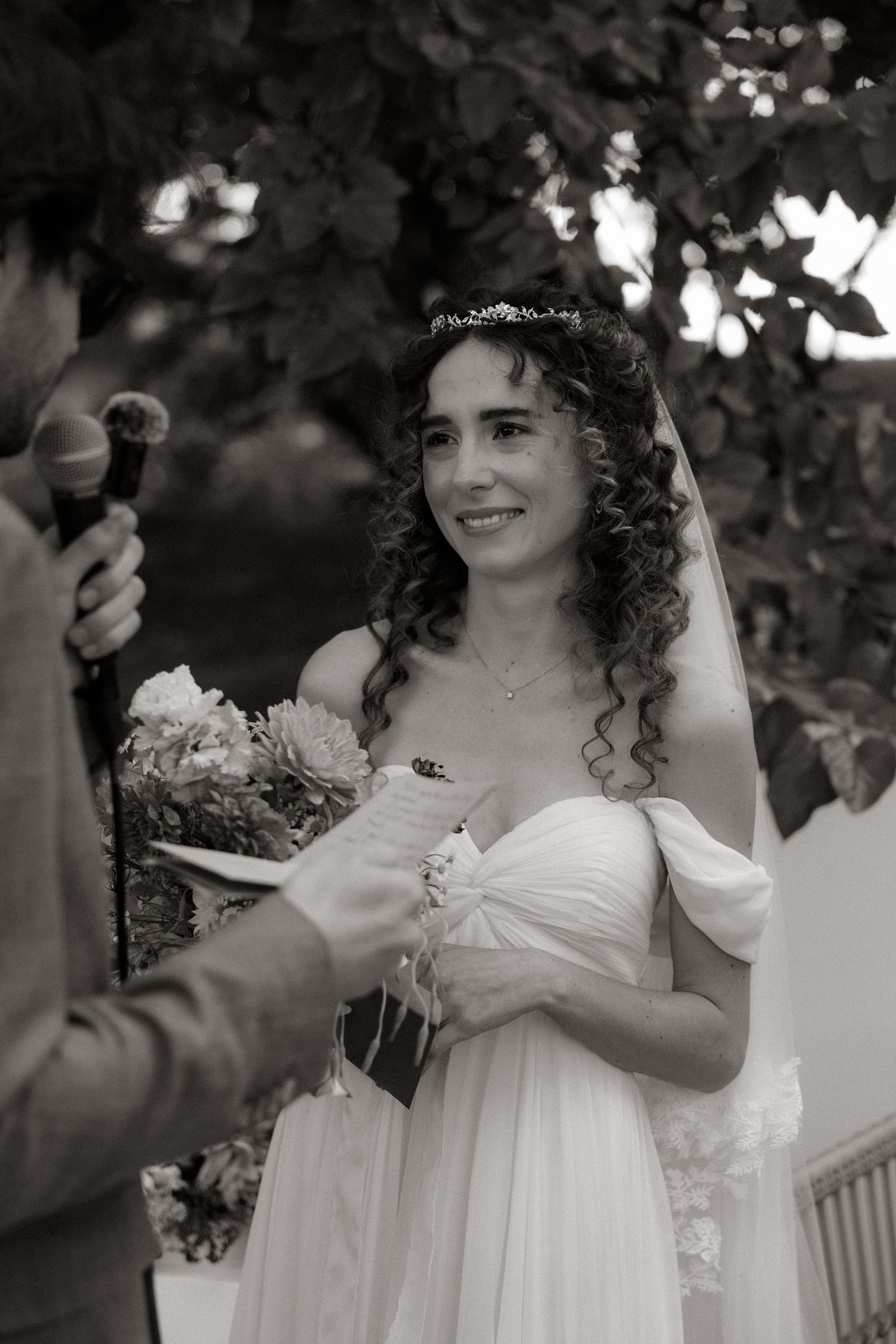 A bride in a flowing white off-shoulder dress holds a bouquet during an outdoor wedding ceremony in black and white.