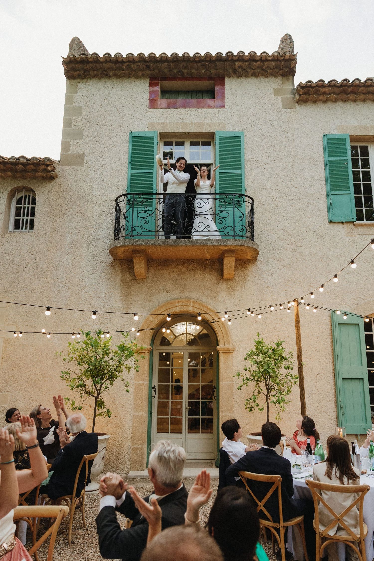 Guests wave to newlyweds on balcony of Mediterranean villa with string lights.
