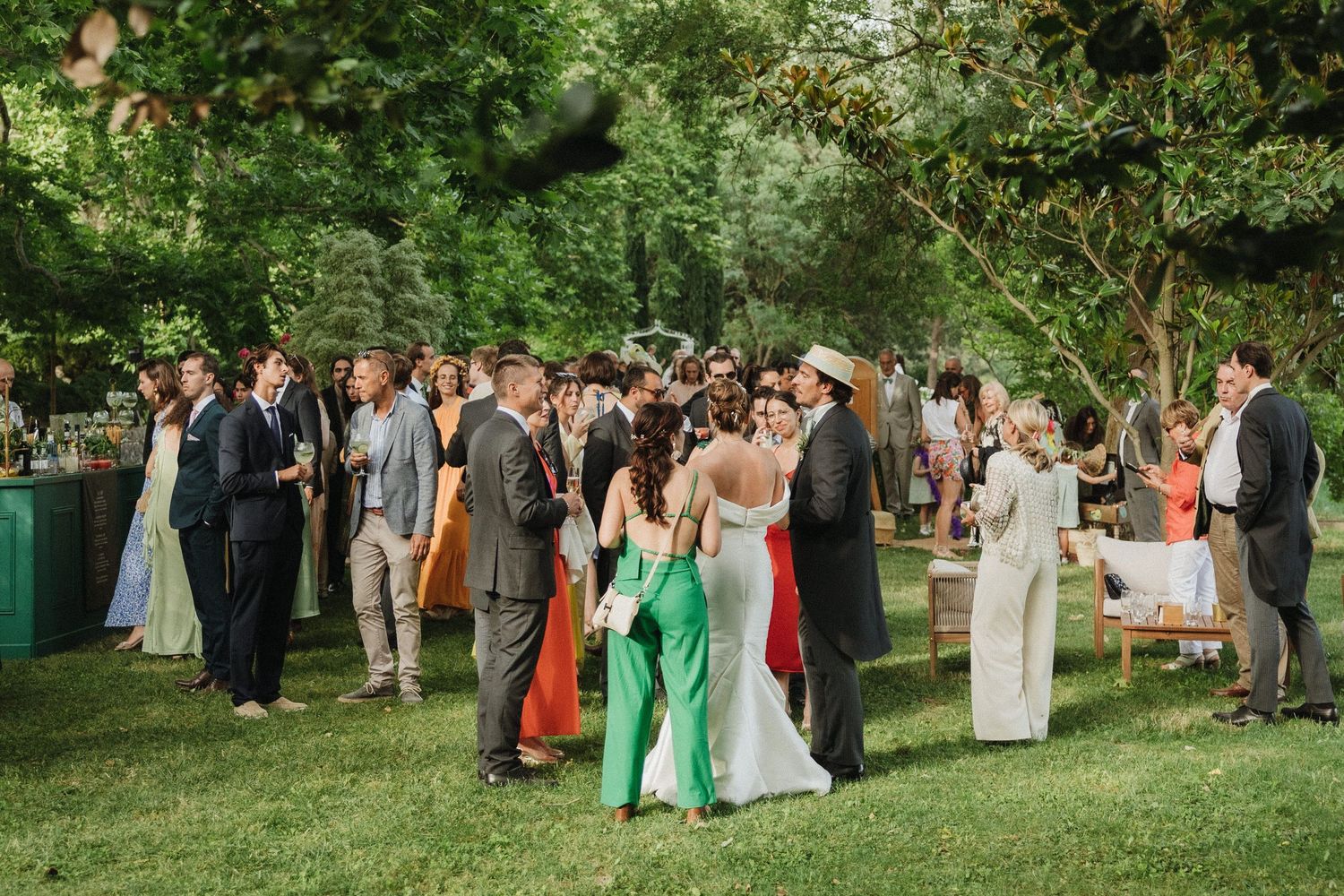 Large group of wedding guests gathered outdoors in formal attire under green trees for a group photo.