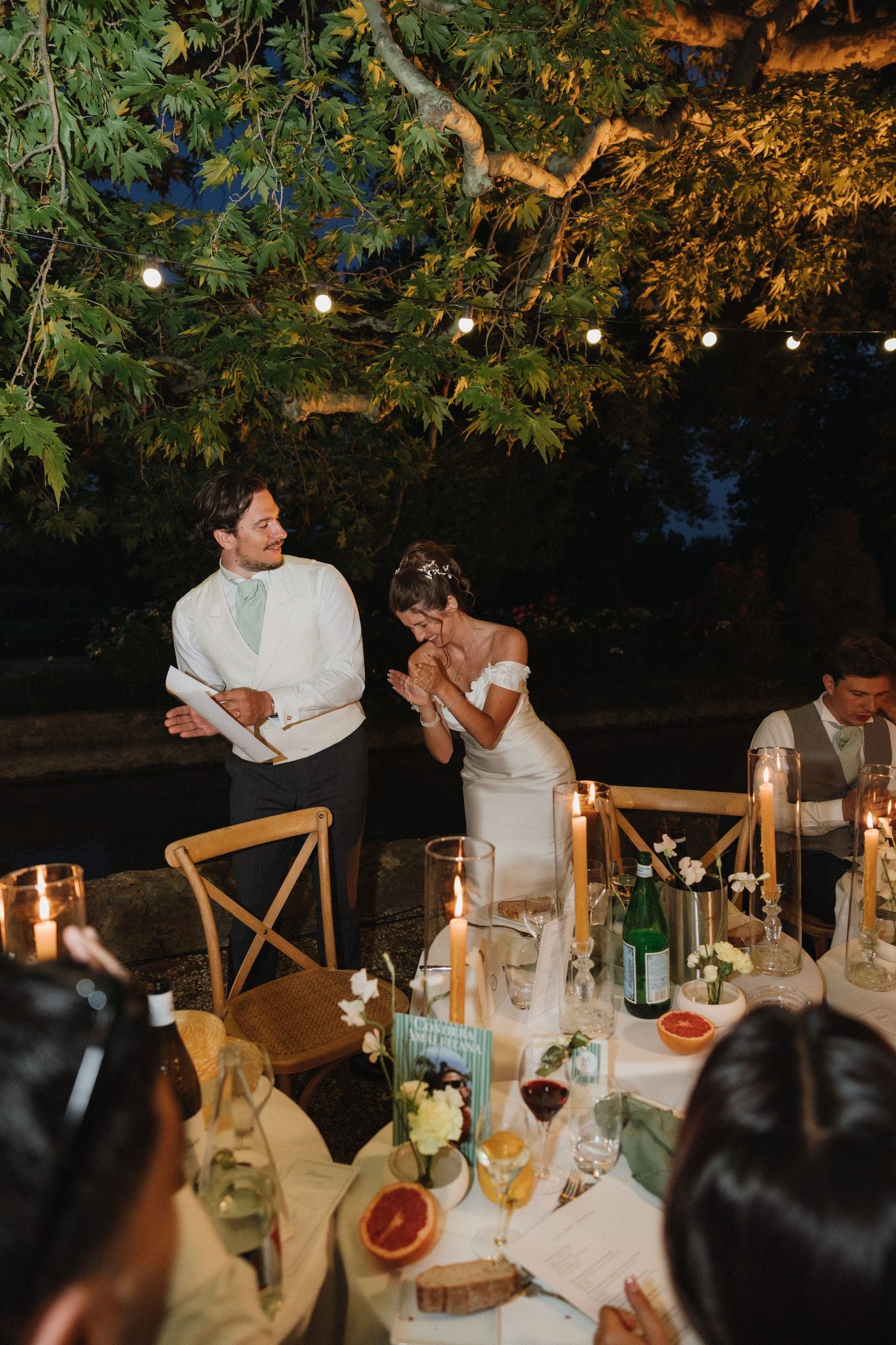 Evening outdoor wedding reception with string lights and table settings under trees.