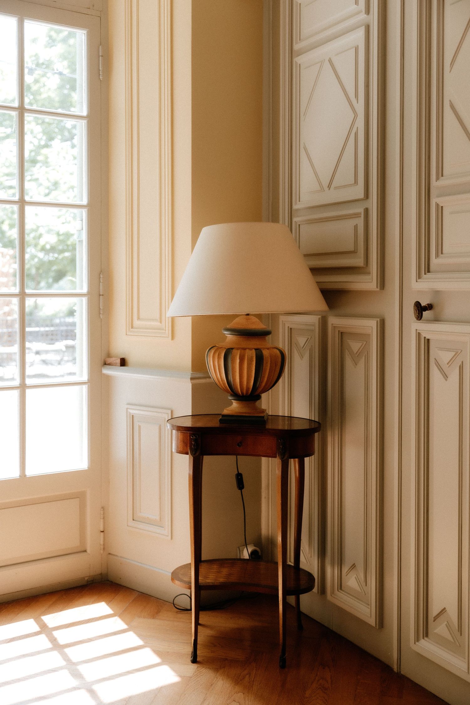 Elegant interior room with white paneled walls, tall windows, and decorative furniture casting dramatic shadows.