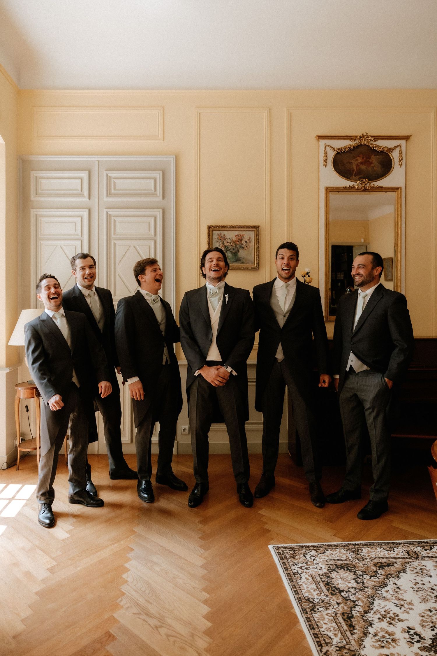 Groomsmen lounging on floor in formal suits showing off colorful socks in elegant room with hardwood floors.