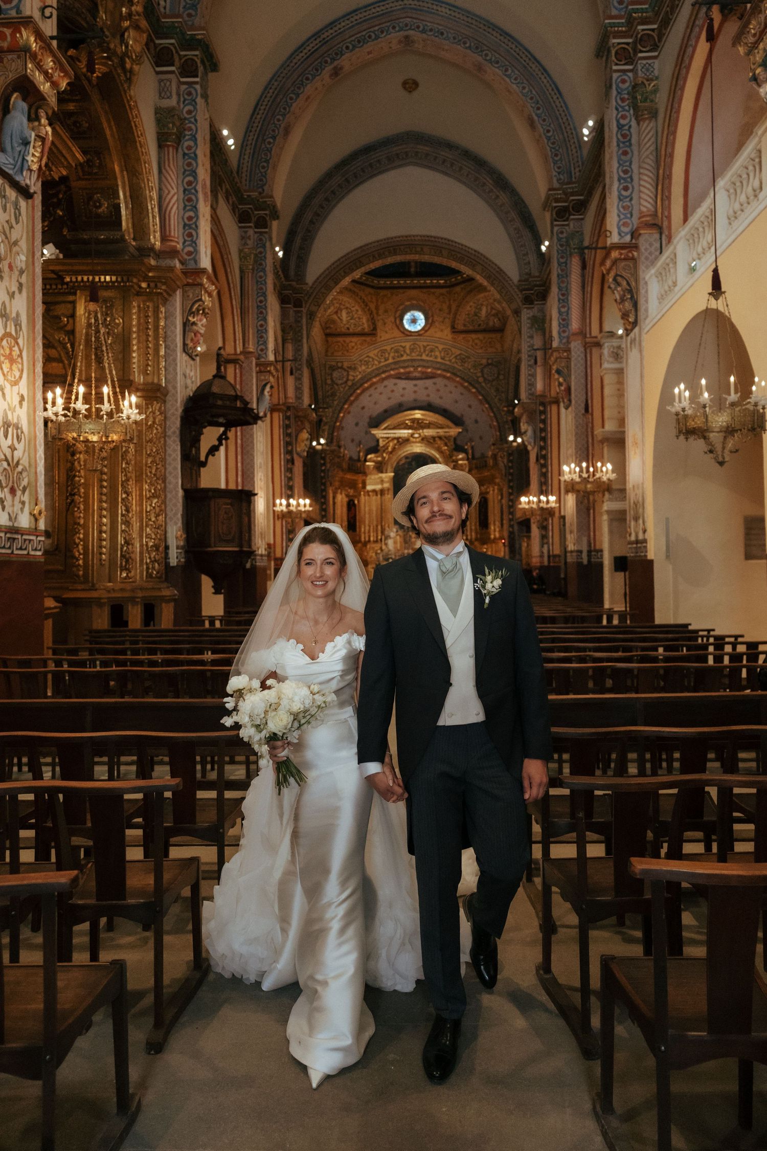 Couple walking down the aisle of a beautiful historic church with vaulted ceilings and chandeliers.