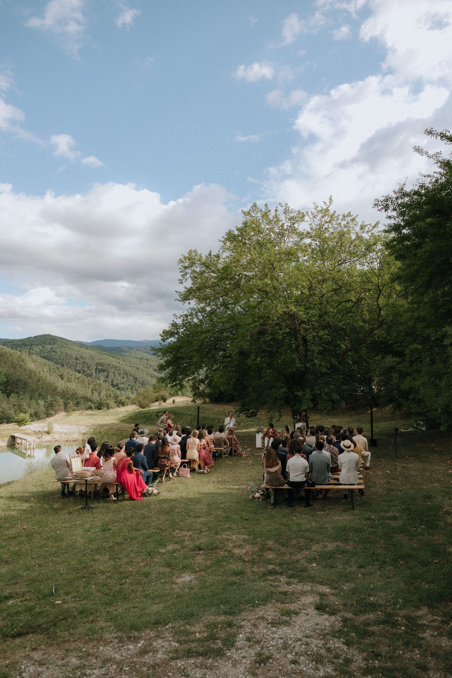 A group gathers for an outdoor wedding ceremony on a grassy hillside with mountain views and scattered trees.