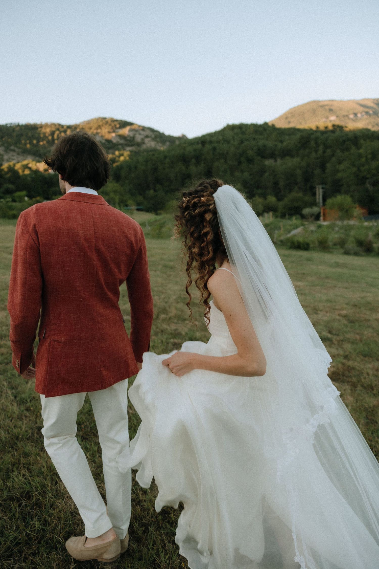 A person in a burgundy jacket and white pants walks alongside someone in a flowing white wedding dress.