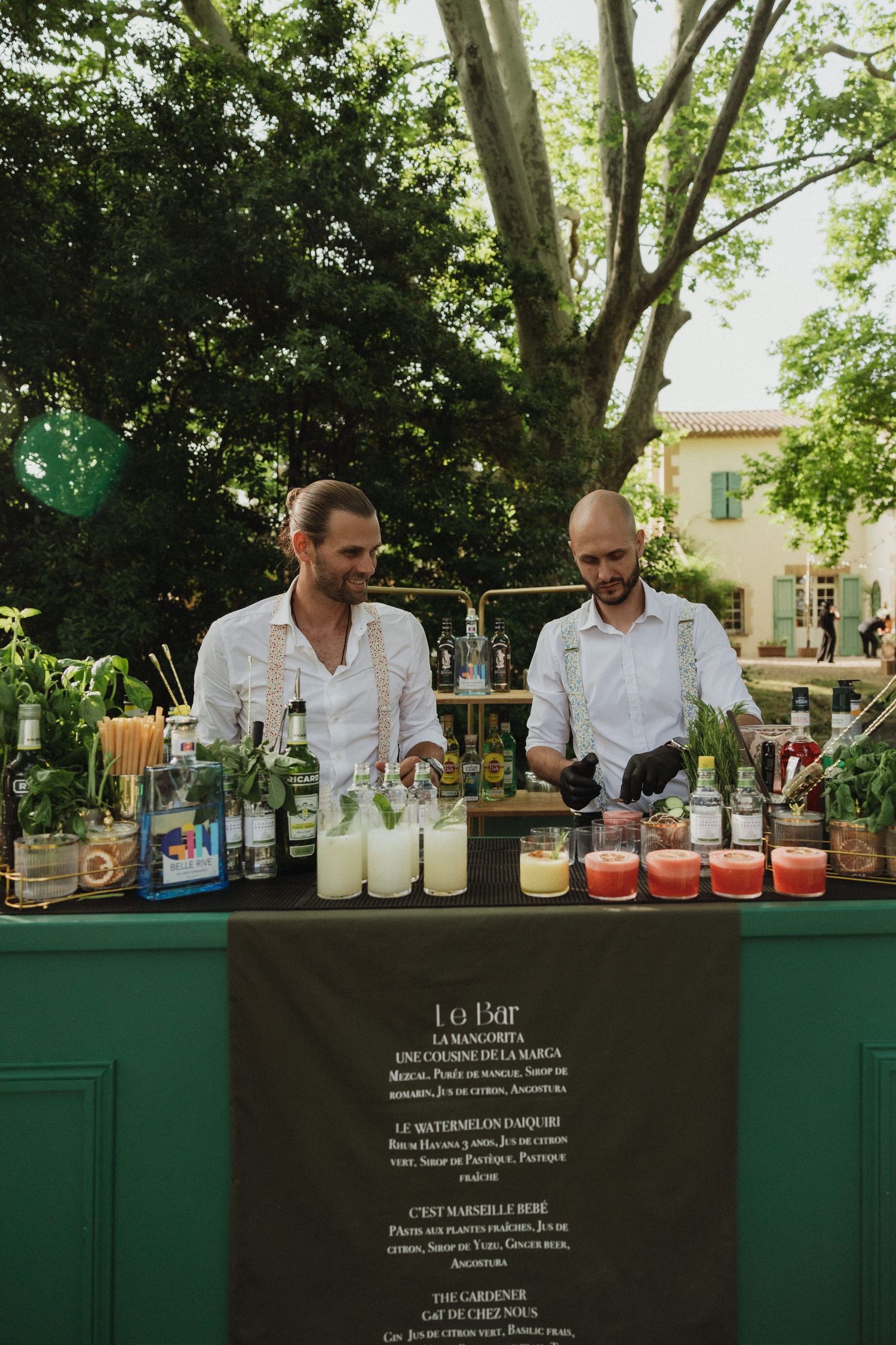 Two bartenders prepare drinks at an outdoor wedding bar with various bottles and mixers.