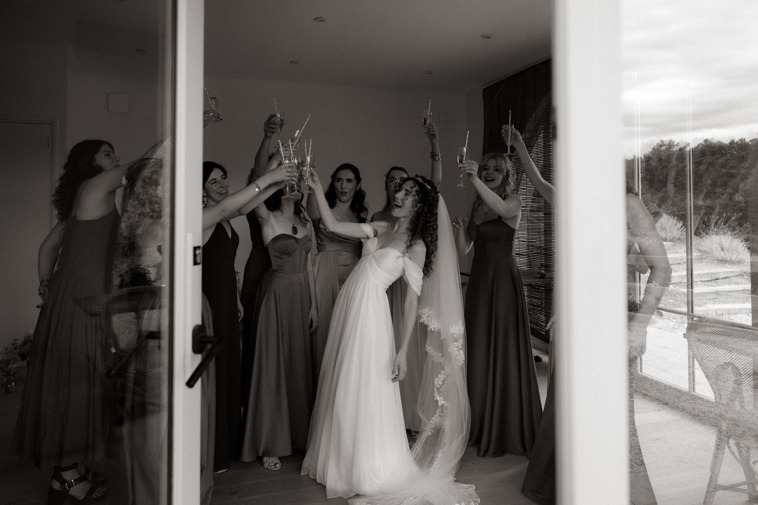 Bridesmaids in floor-length dresses celebrate joyfully with a bride in white during a candid black and white moment.
