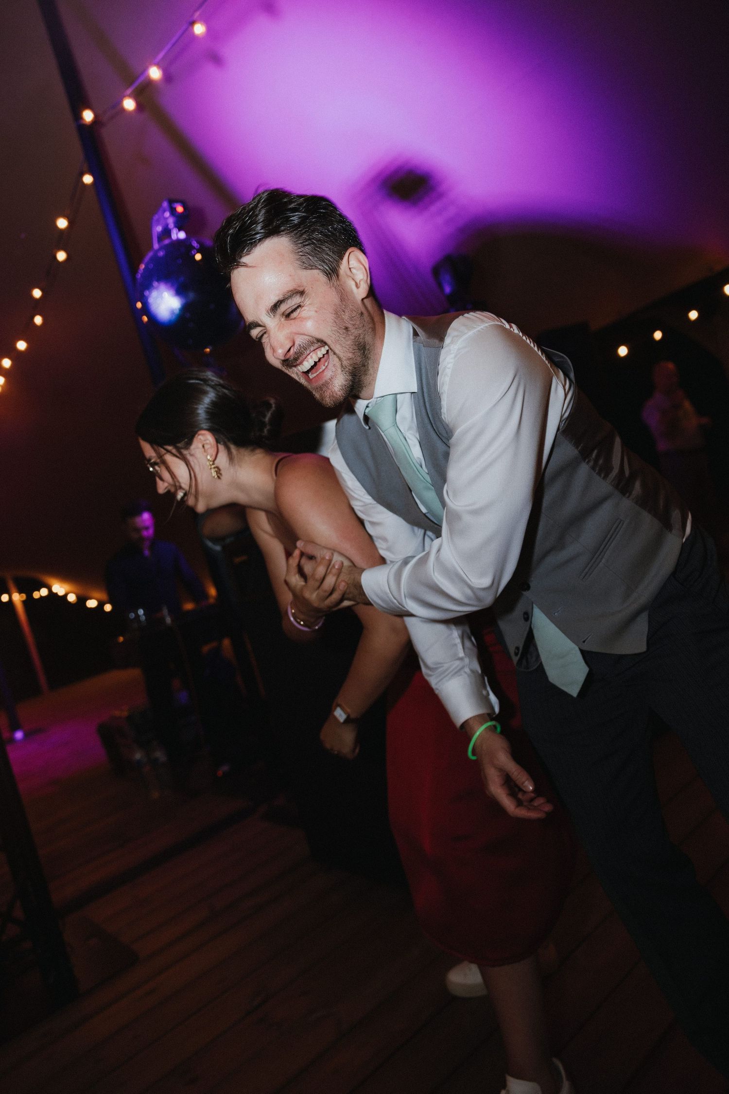 Energetic dancing and laughter at wedding reception with purple mood lighting.
