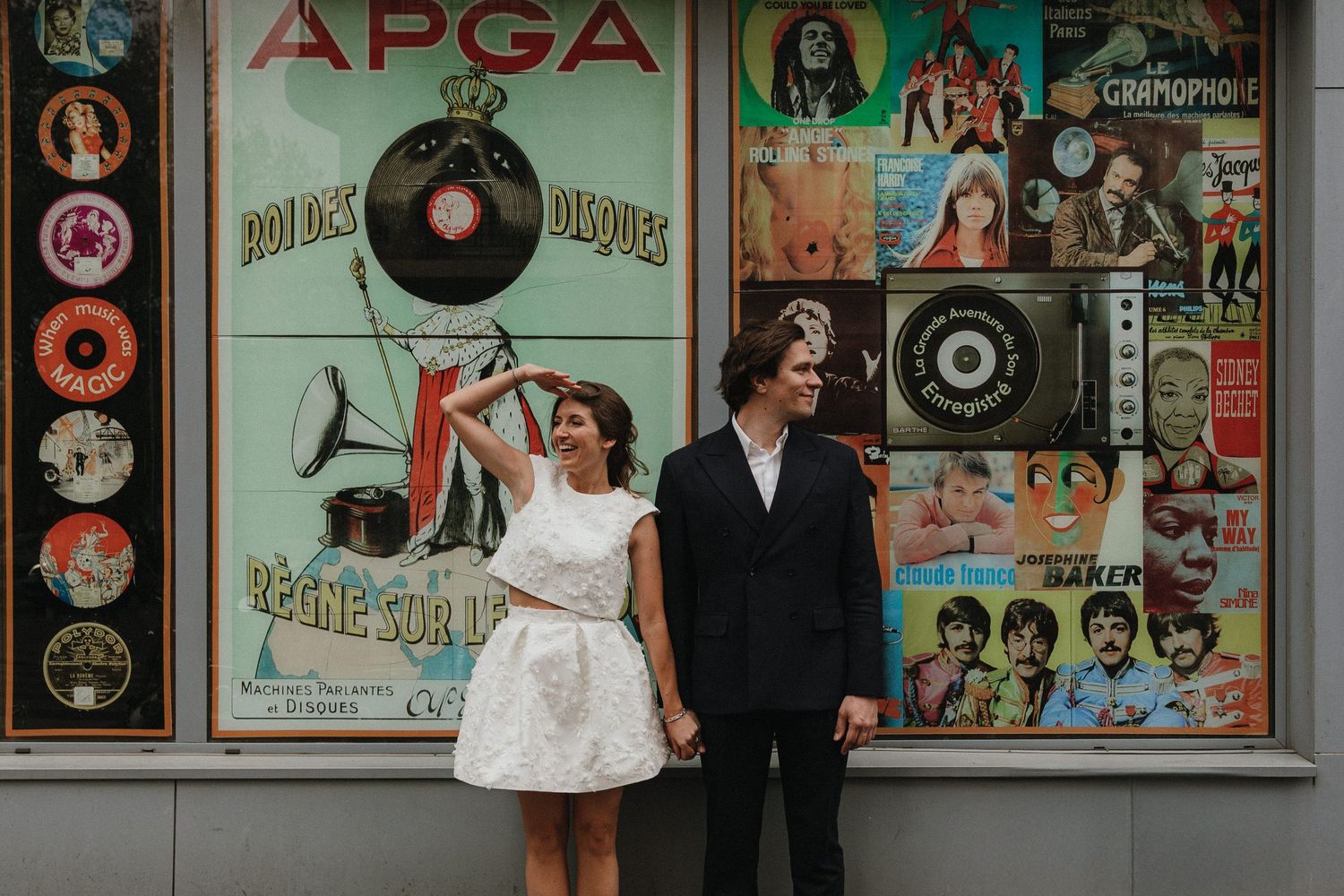 A couple poses together against a wall decorated with vintage vinyl records and retro music posters.