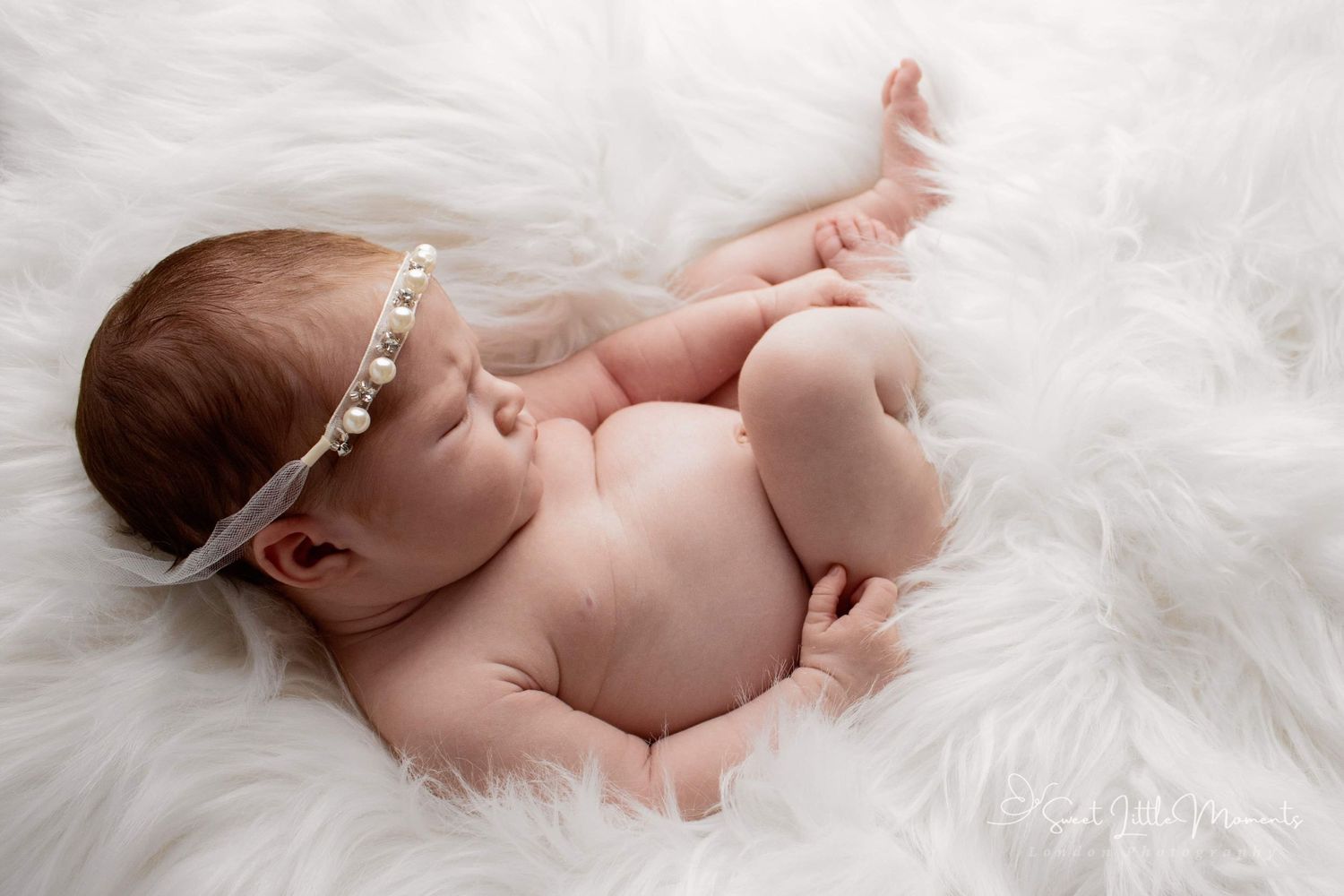 Newborn wearing delicate headband lying on white furry blanket during photo session.