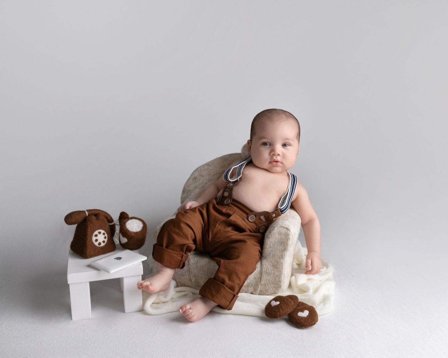 Baby in brown overalls sitting on white bench with teddy bear props.