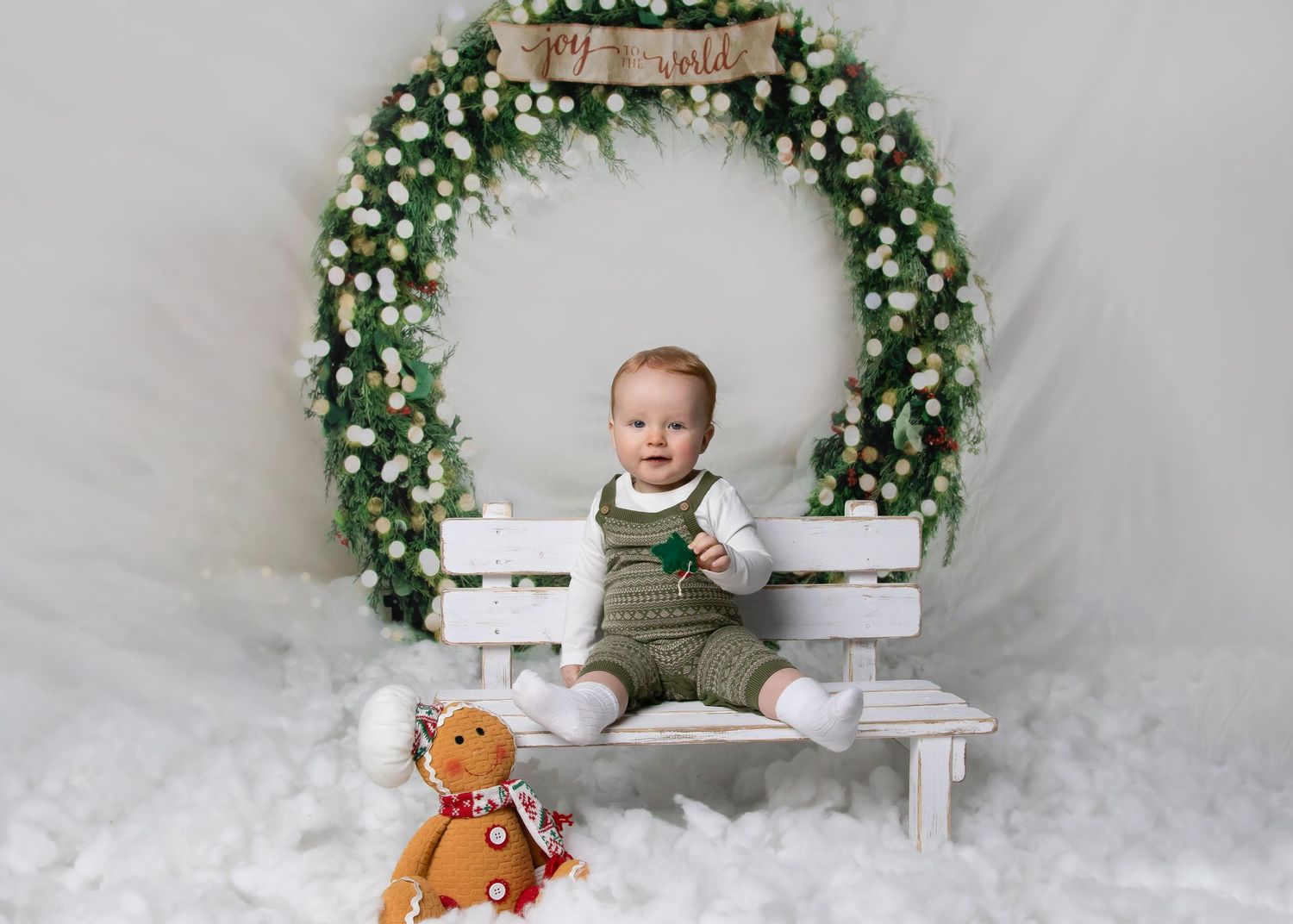 Baby sitting on white bench with floral arch and teddy bear in winter themed photoshoot.